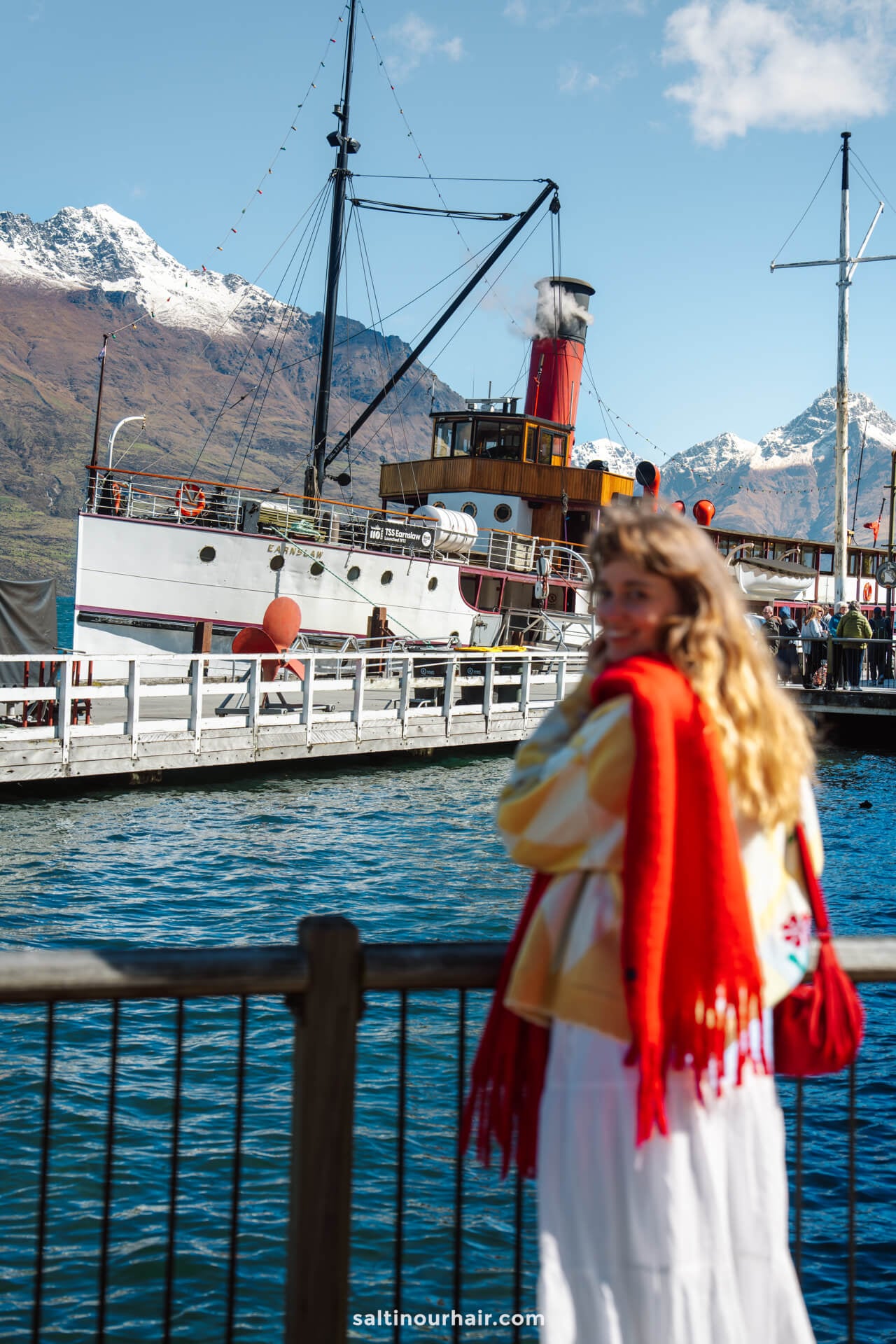 A woman in a red shawl stands by a railing overlooking the docked TSS Earnslaw steamship, with mountains and blue sky in the background&mdash;one of the best things to do in Queenstown, new zealand