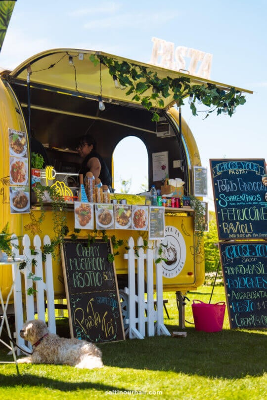 A yellow food truck selling pasta dishes is parked on grass with menus displayed outside. A person works inside, and a dog lies out front&mdash;one of the must-try things to do in Queenstown for food lovers.
