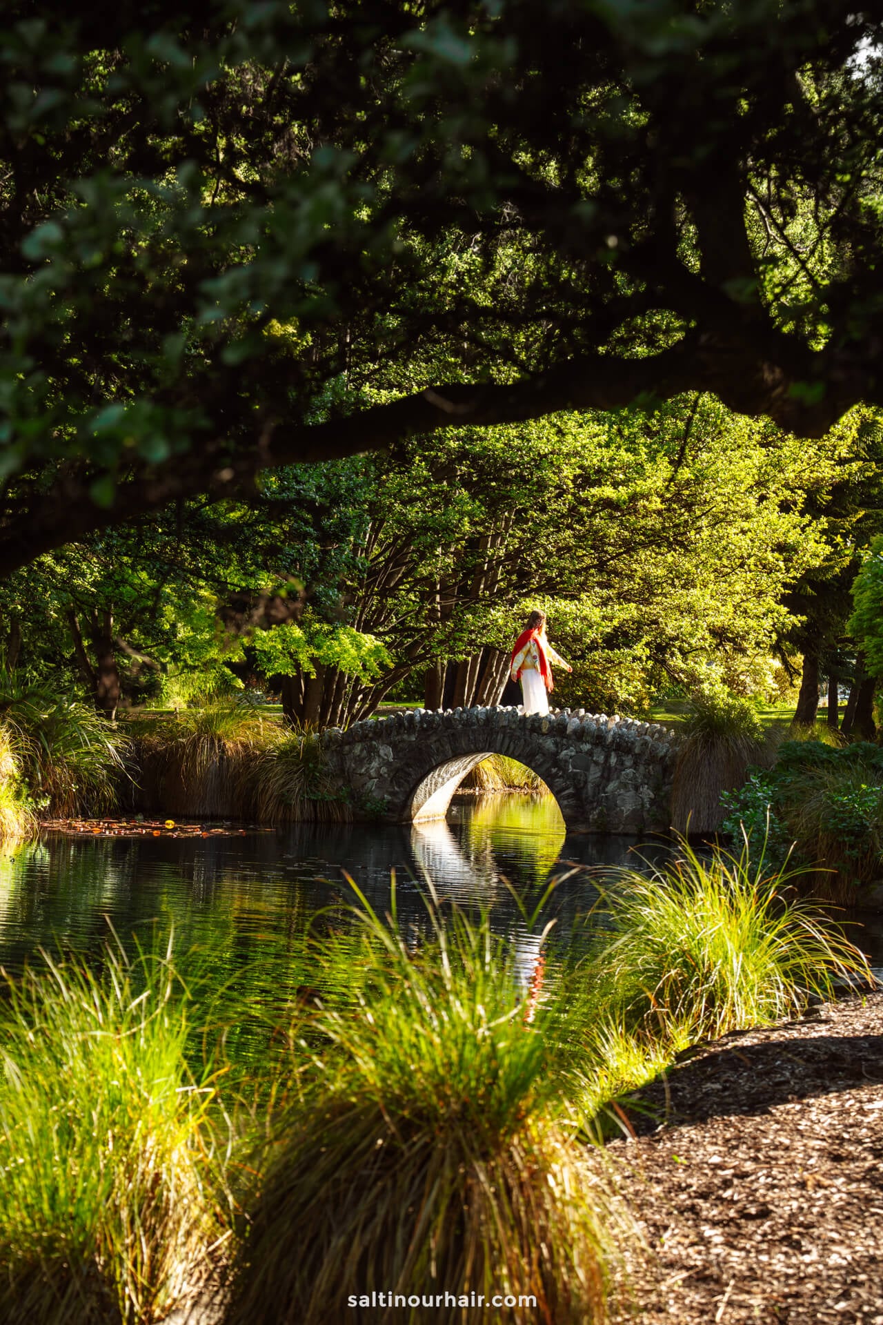 A woman in a white dress and red scarf stands on a small stone bridge over a pond in queenstown gardens, new zealand