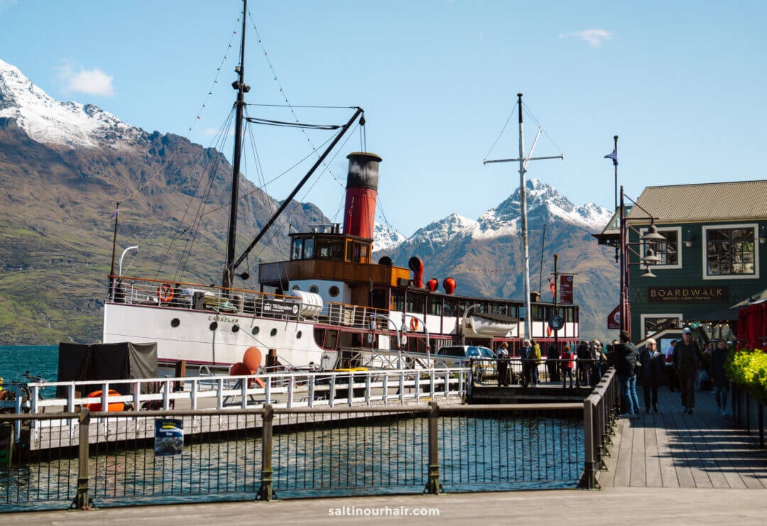 A vintage steamship is docked at a wooden pier in queenstown new zealand, with mountains and snow-capped peaks in the background