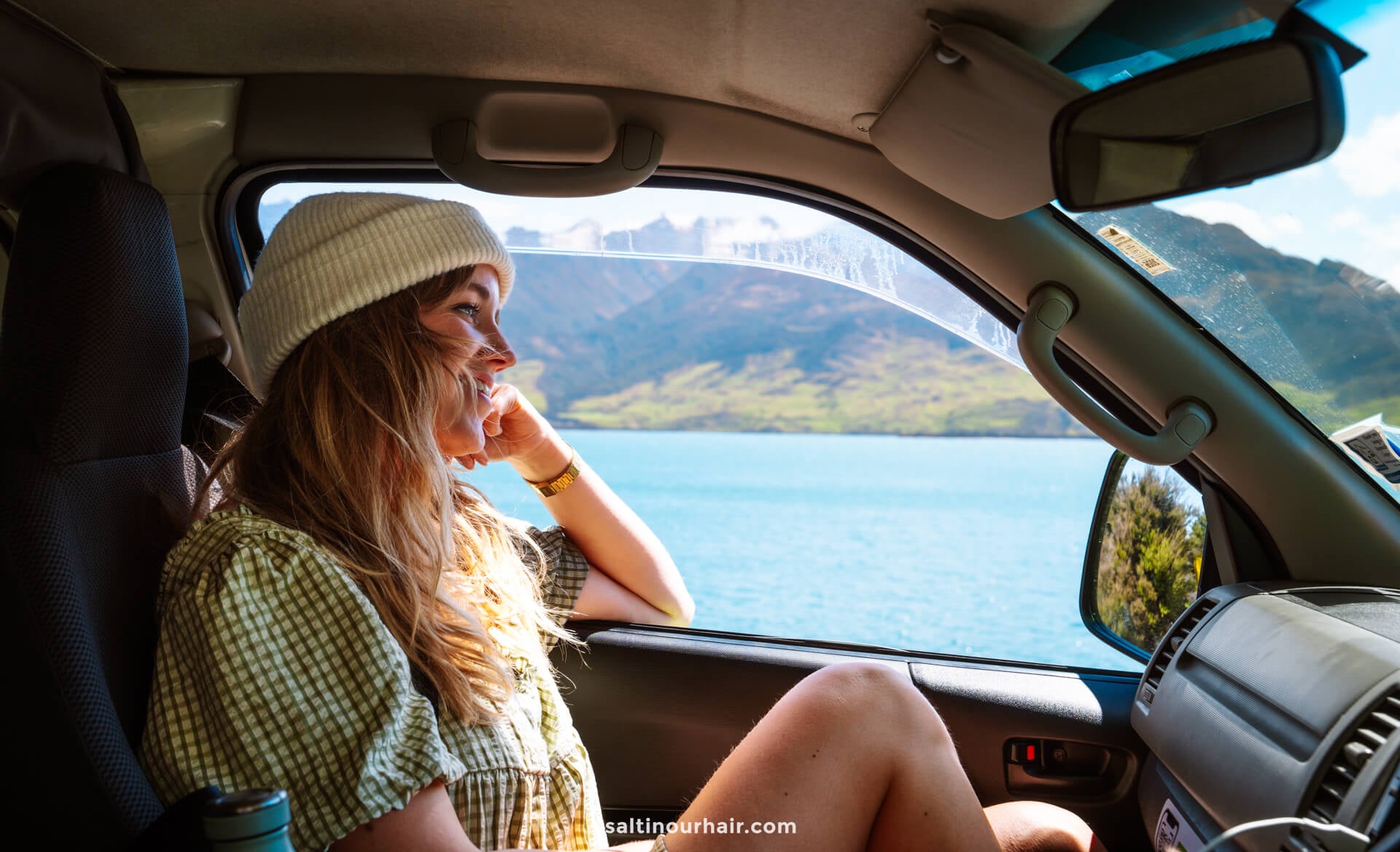 A woman wearing a white beanie sits in the passenger seat of a van, gazing out the window at a lake and mountains&mdash;soaking in the bright sky and dreaming of things to do in Queenstown, new zealand