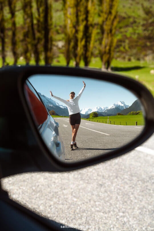A person in athletic wear poses with arms raised on a road, reflected in a car side mirror, with mountains and trees in the background&mdash;capturing the adventurous spirit of things to do Queenstown, new zealand