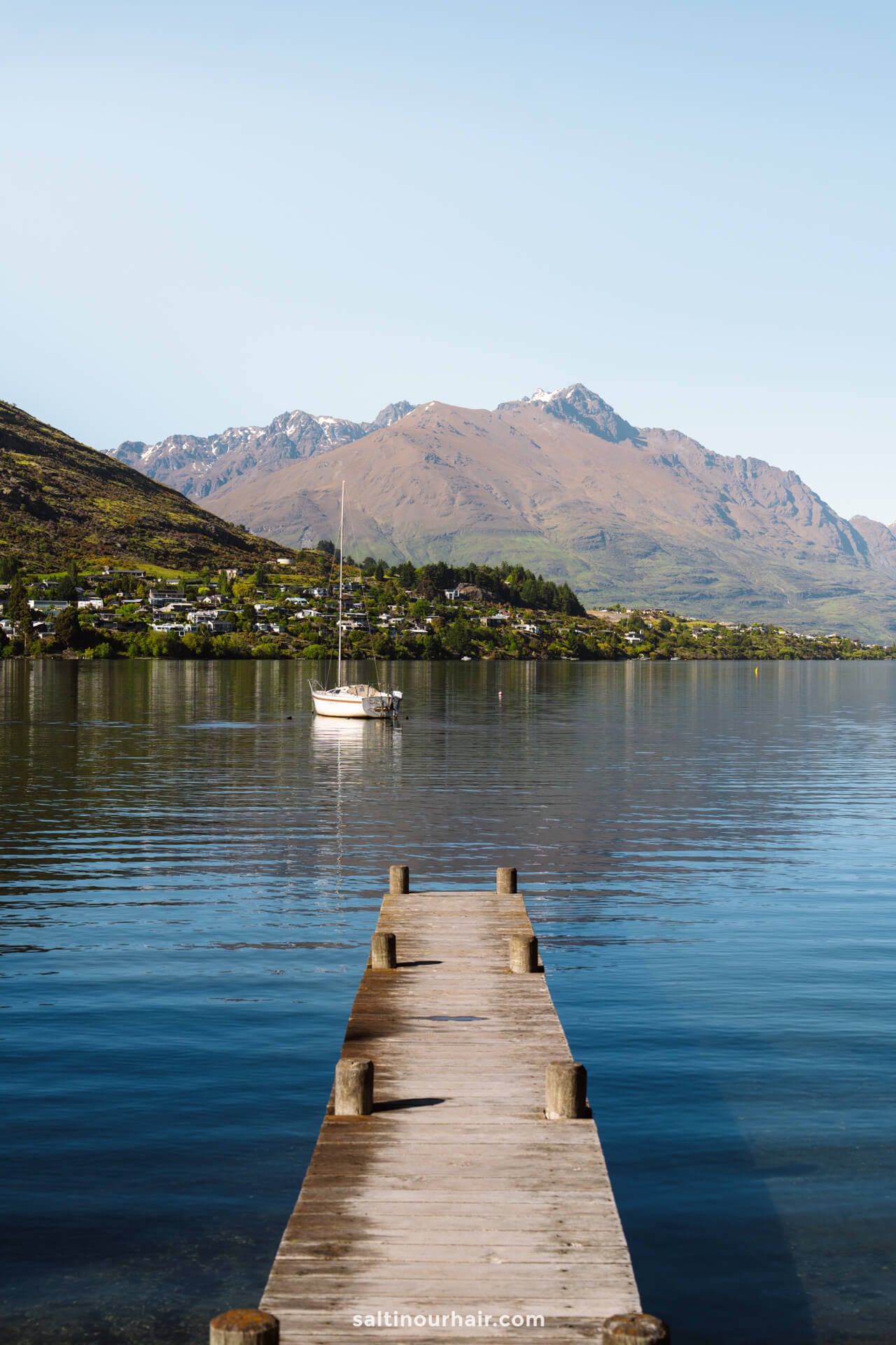 A wooden dock stretches into a calm lake with a sailboat anchored nearby; mountains and houses are visible in the background under a clear sky&mdash;capturing the serene beauty and things to do Queenstown, new zealand has to offer.