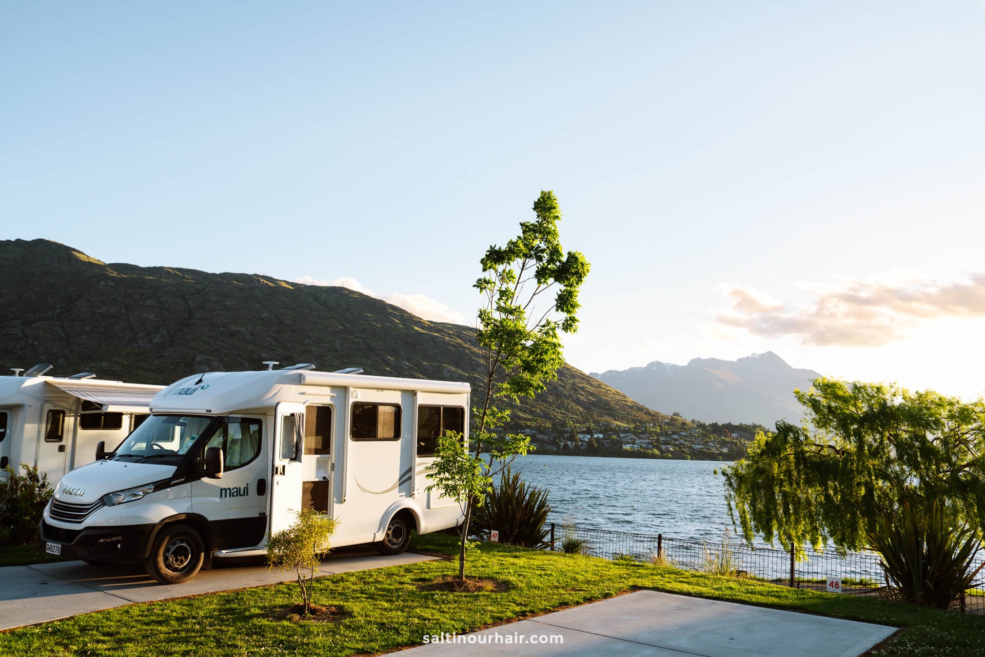 A white camper van is parked by a lakeside at Driftview Campsite with mountains, trees, and houses in the background under a clear sky&mdash;perfect for exploring the many things to do Queenstown has to offer.