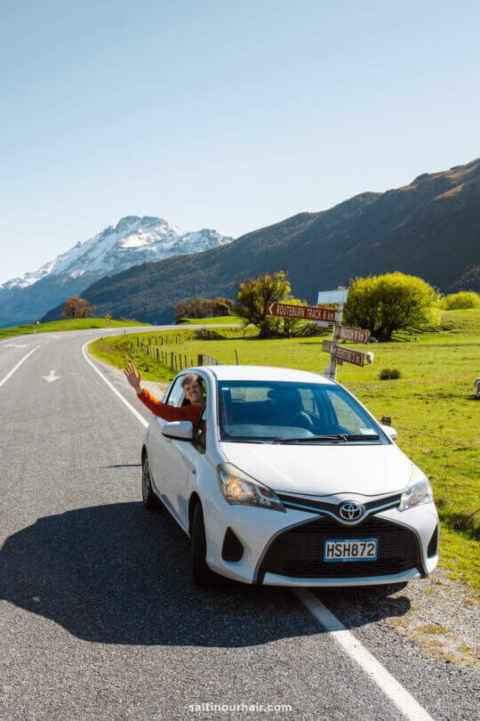 A person waves from the driver&rsquo;s seat of a white Toyota parked on a rural road near Queenstown, with mountains and a signpost in the background&mdash;perfect for those exploring things to do in Queenstown, new zealand