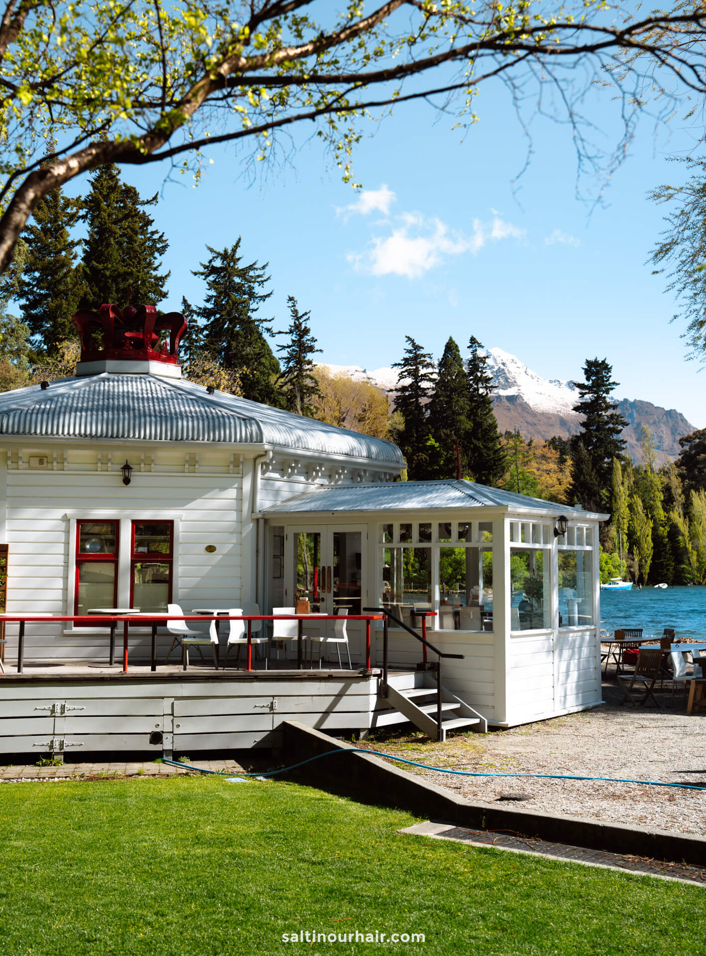 A white lakeside building with a metal roof and outdoor seating, surrounded by trees, offers a serene spot to relax while enjoying one of the many things to do Queenstown has to offer, with mountains and a blue sky in the background.