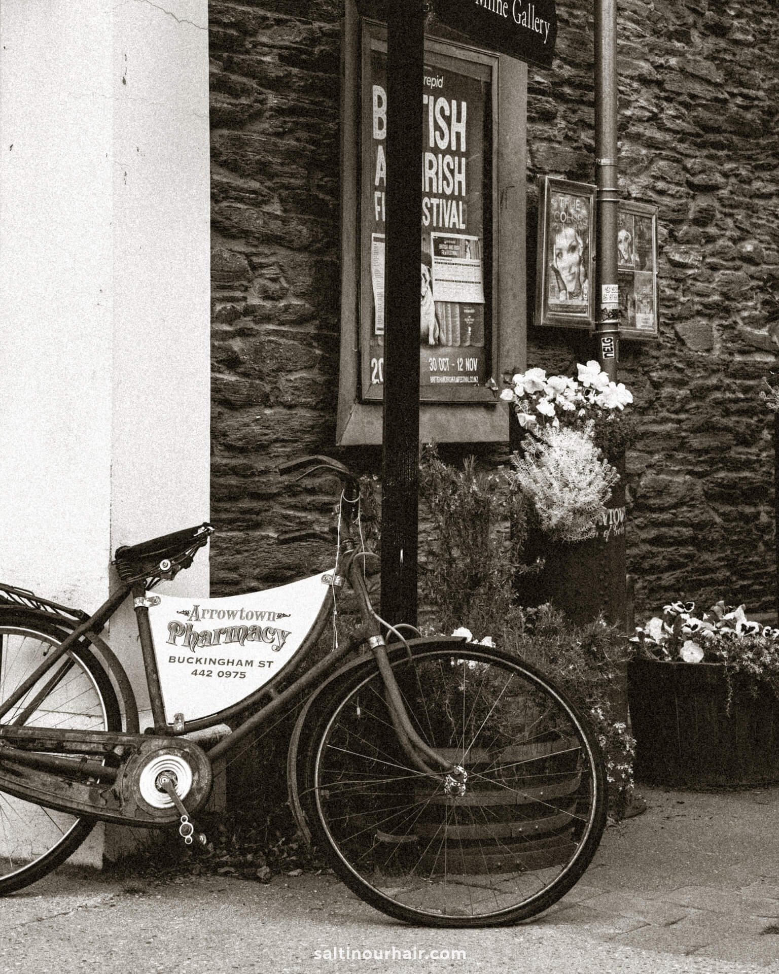 A vintage bicycle leans against a wall outside a shop in Arrowtown, New Zealand, with flower pots and festival posters visible in the background.