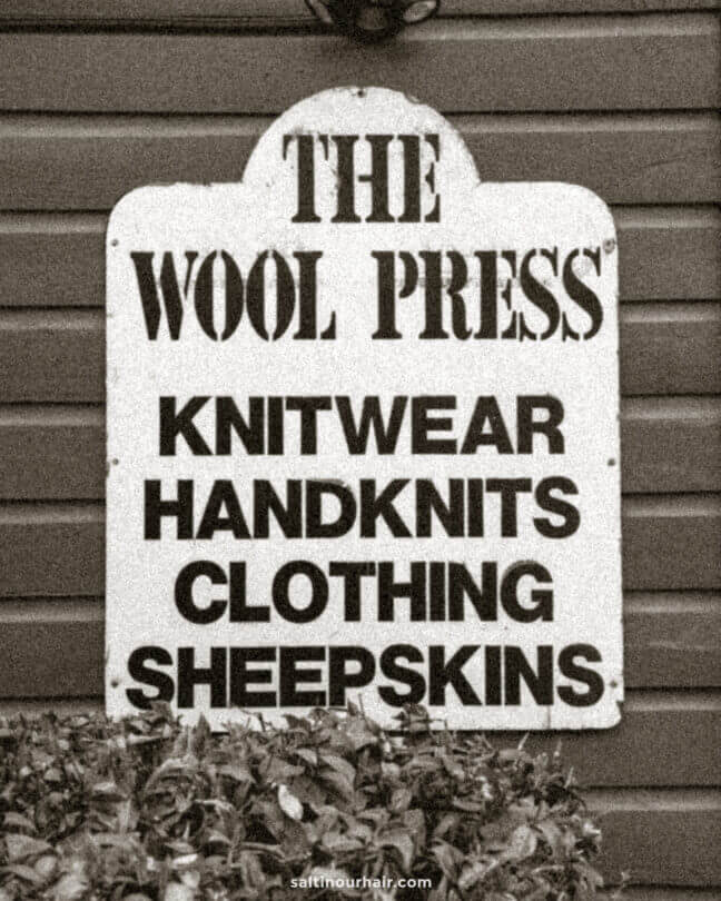 A sign in Arrowtown, New Zealand reads: THE WOOL PRESS KNITWEAR HANDKNITS CLOTHING SHEEPSKINS and is mounted on a wooden wall above some foliage.