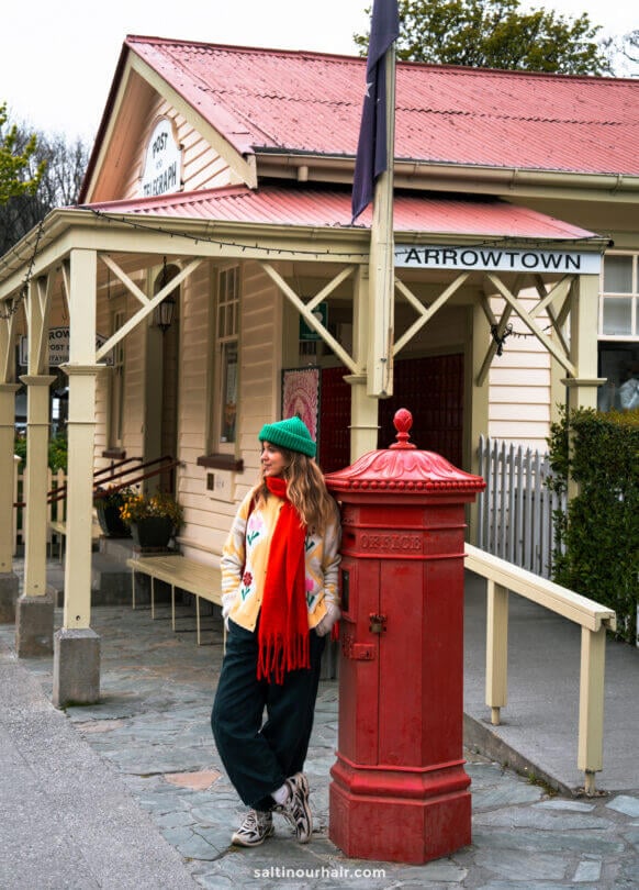 A woman in colorful winter clothes stands by a red post box in front of the historic Arrowtown Post and Telegraph building in Arrowtown, New Zealand.