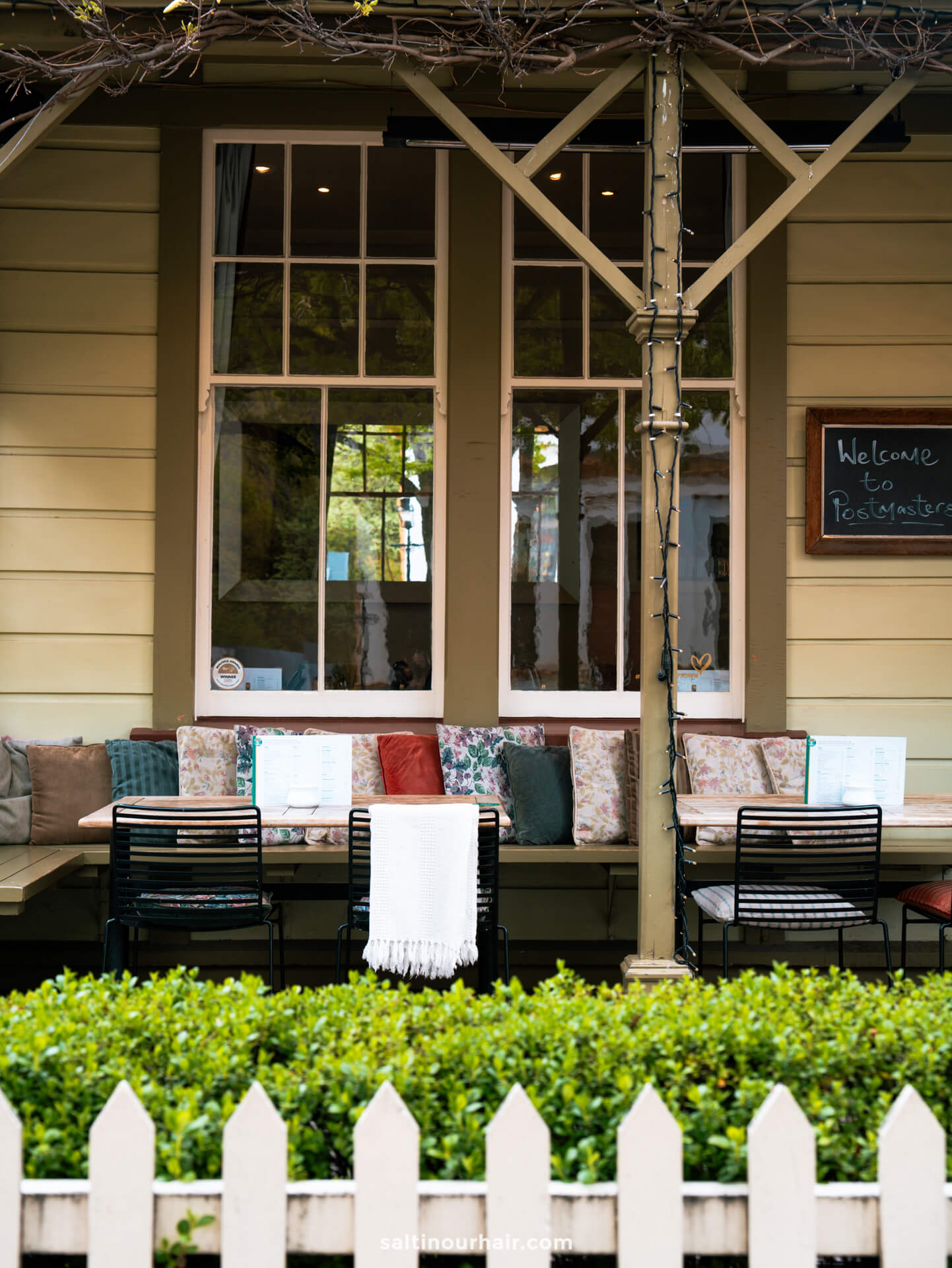 Outdoor cafe seating area with cushioned benches, a table set with menus, black chairs, and a Welcome to Toastmasters sign on the wall, nestled behind a white picket fence and greenery in charming Arrowtown, New Zealand.
