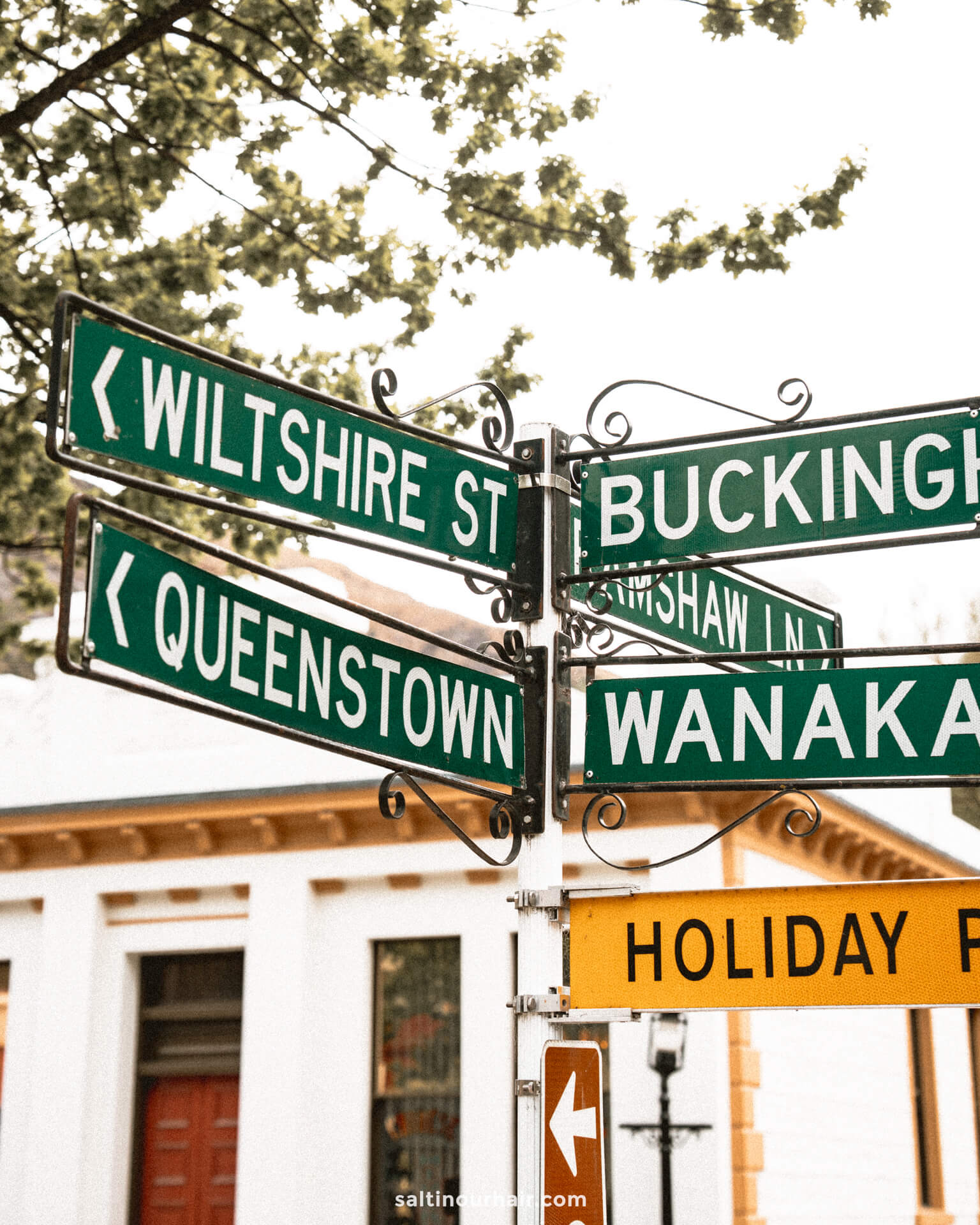 Street signs at an intersection point to Wiltshire St, Queenstown, Arrowtown, Buckingham, and Wanaka, with a yellow Holiday Park sign below. Trees and a building are in the background.