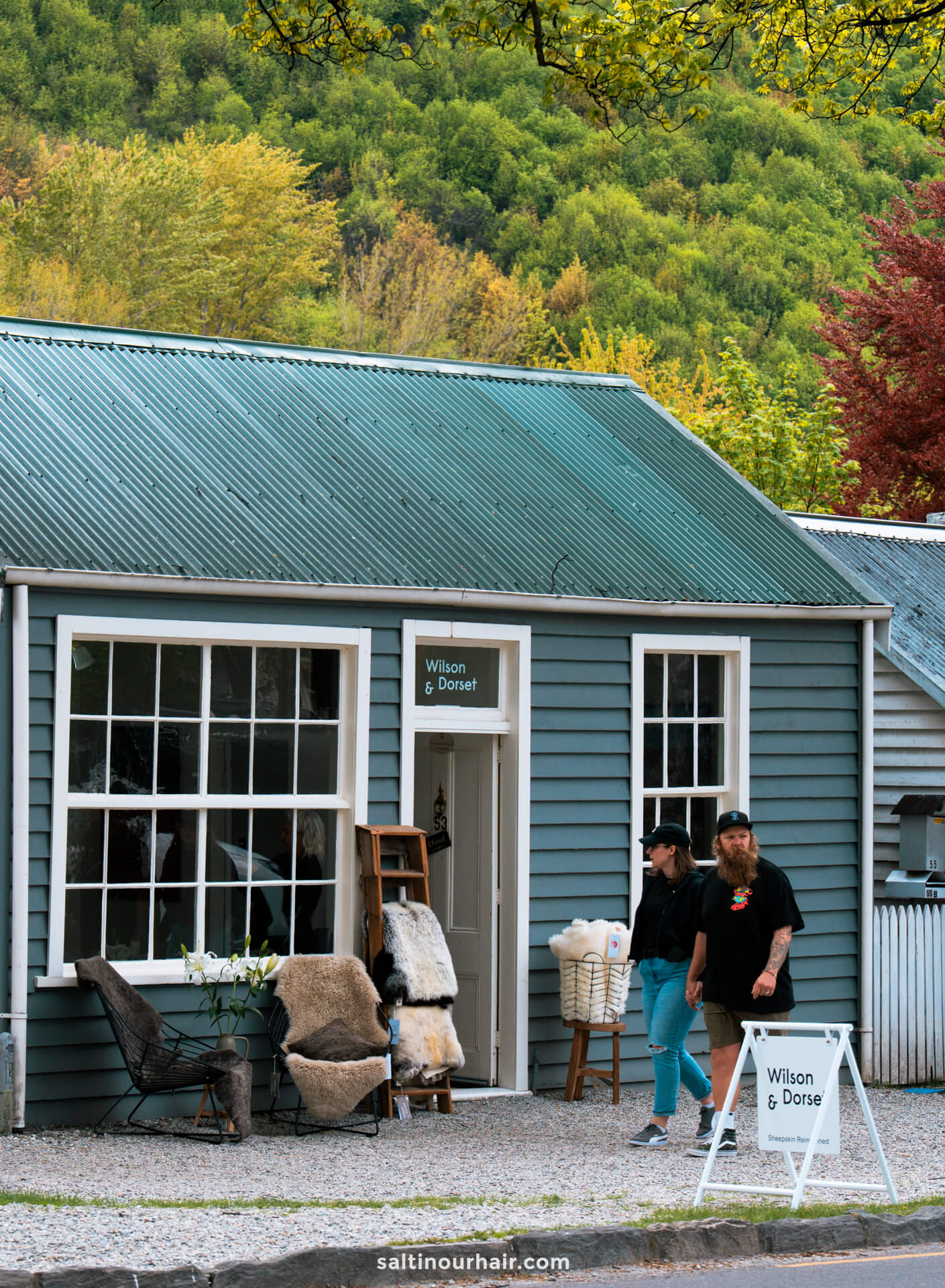 Two people stand outside Wilson &amp; Dorset, a small shop with blue siding and large windows in Arrowtown, New Zealand, displaying rugs and chairs on a tree-lined street.