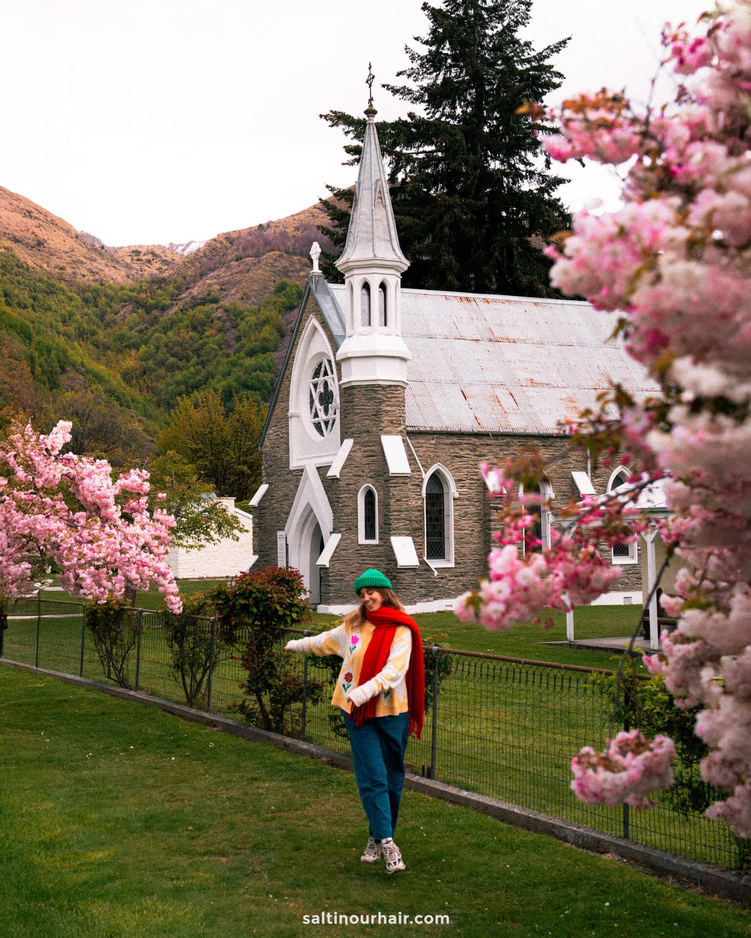 A person poses on a grassy lawn lined with blooming pink trees in front of a historic stone church with a white steeple, surrounded by mountains in Arrowtown, New Zealand.