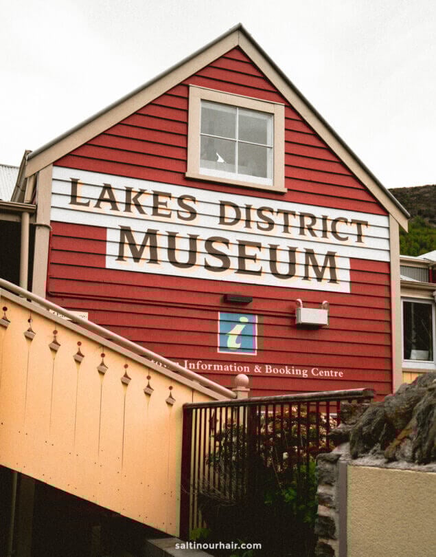 Red building with white trim labeled Lakes District Museum above a visitor information sign, with a staircase and railing in the foreground, located in charming Arrowtown, New Zealand.