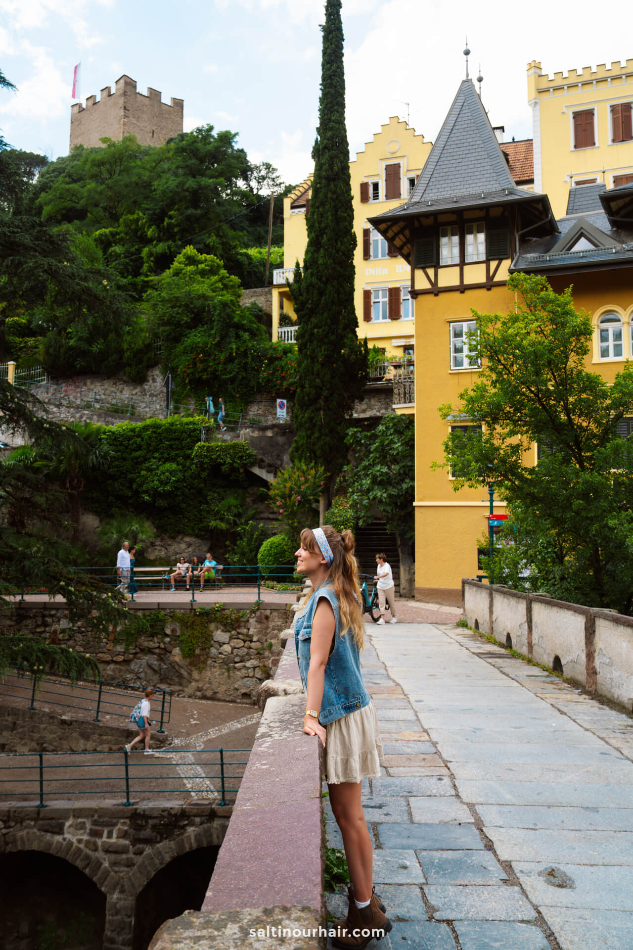 A woman stands on a stone bridge in Merano, Dolomites, Italy, overlooking a historic area with yellow buildings and a castle perched on a hill in the background.