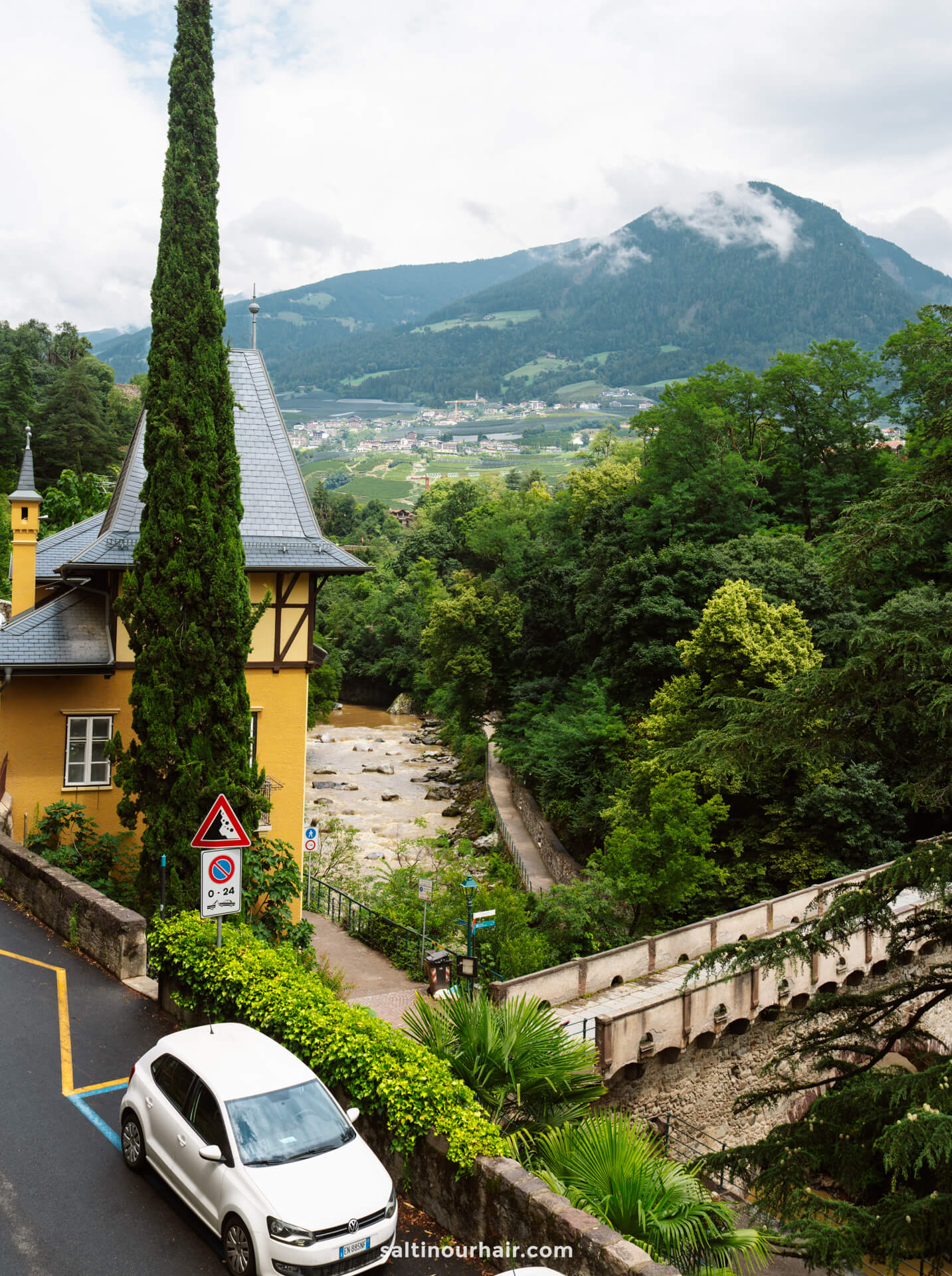 A white car is parked beside a yellow house with a tall cypress tree in Merano Dolomites Italy. 