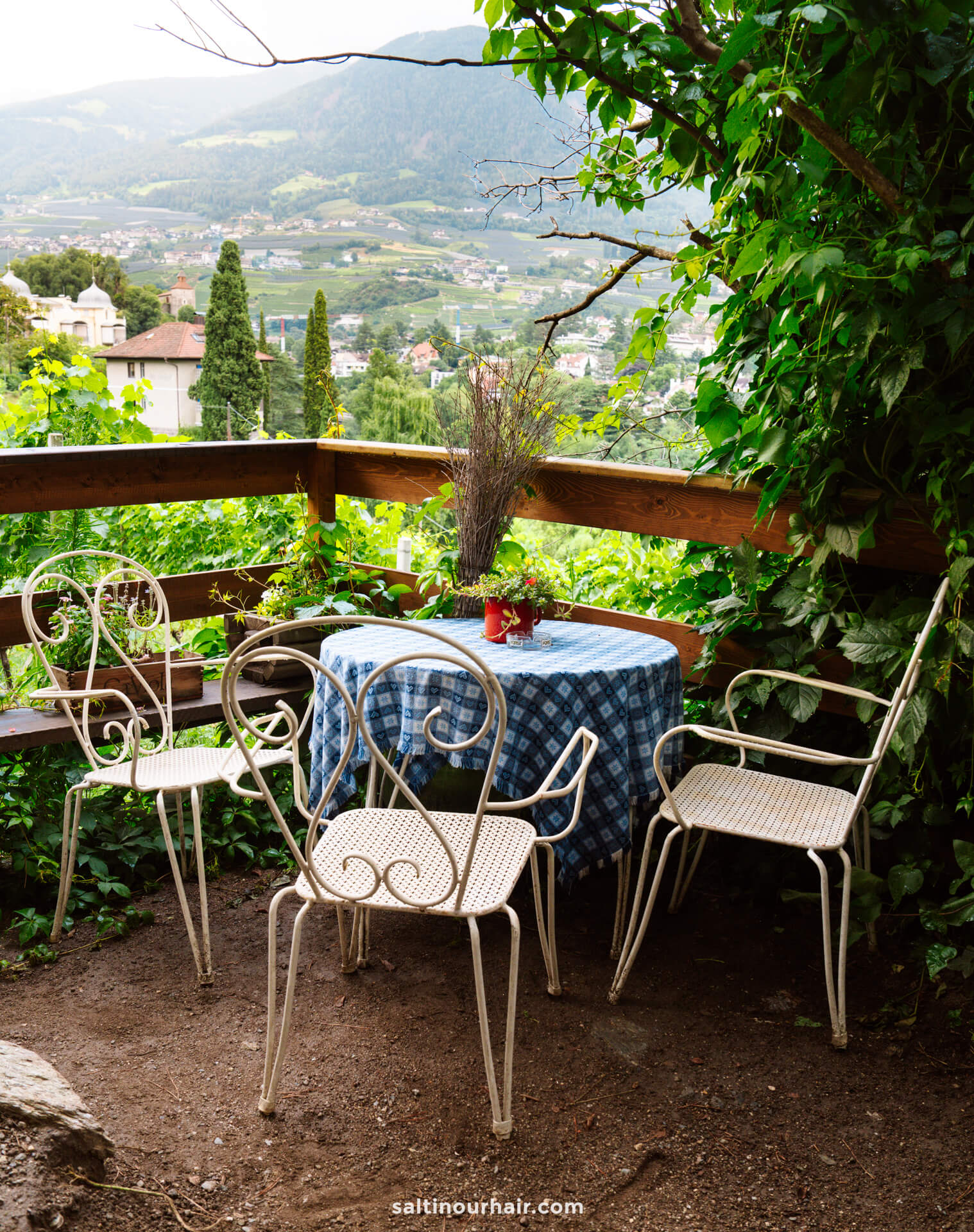 A small outdoor table with a blue checkered tablecloth and three metal chairs sits on a patio in Merano Dolomites, surrounded by greenery and overlooking a lush valley.