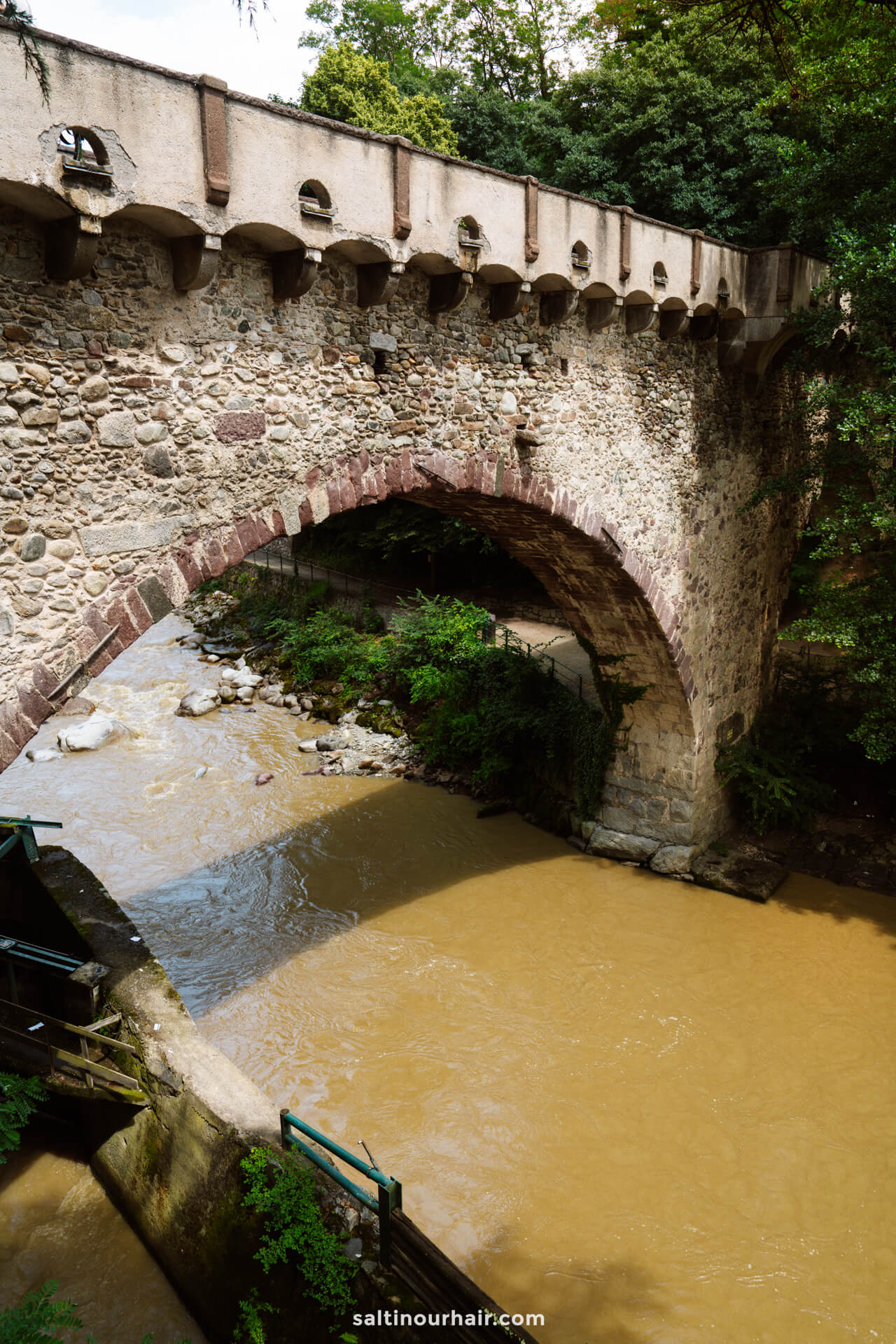 A stone arch bridge with a fortified wall crosses over a muddy river  in the charming town of Merano Dolomites Italy.
