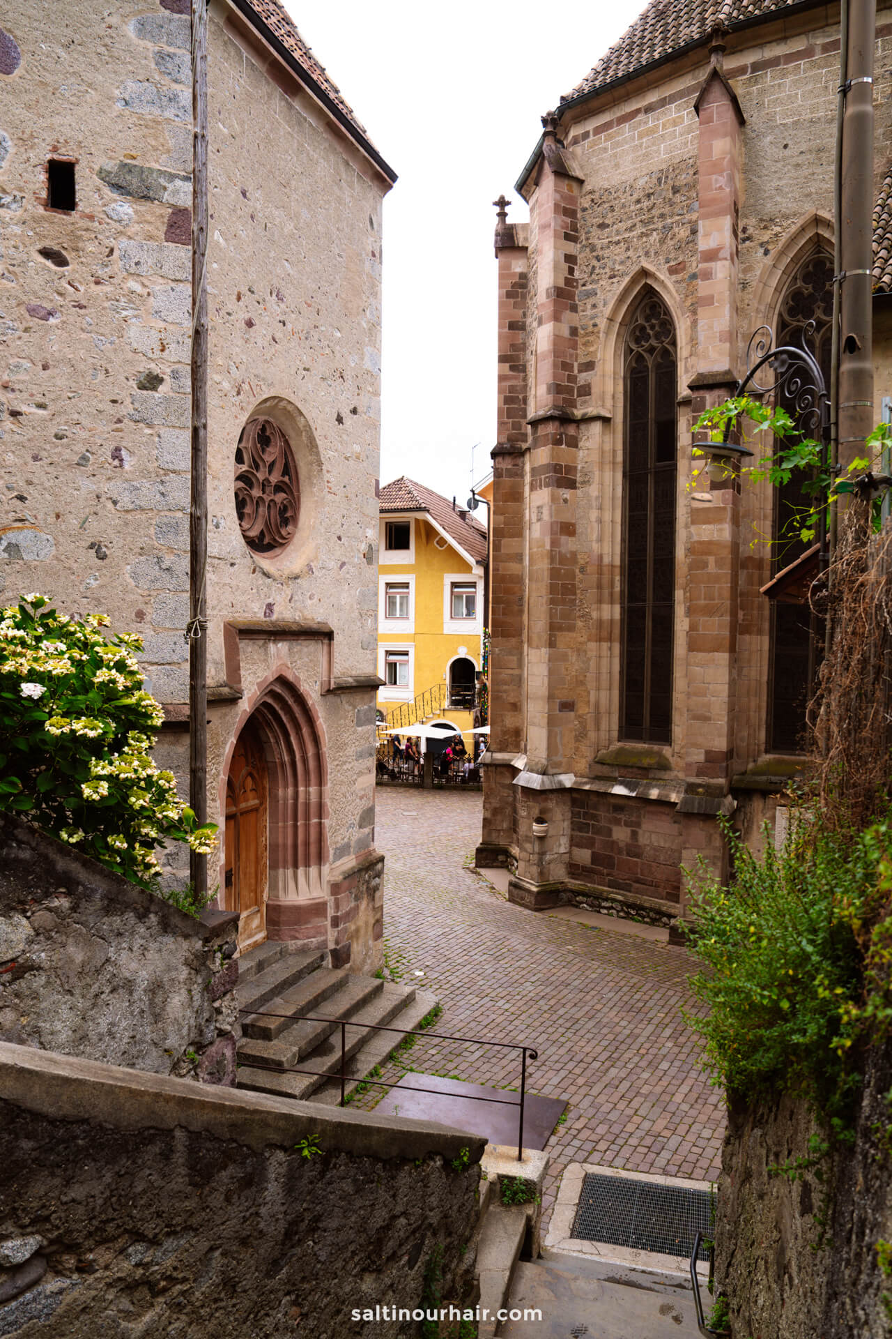 Stone church buildings and a narrow cobblestone street in Merano Dolomites Italy, leading to a small plaza with yellow buildings and people sitting in an outdoor cafe.