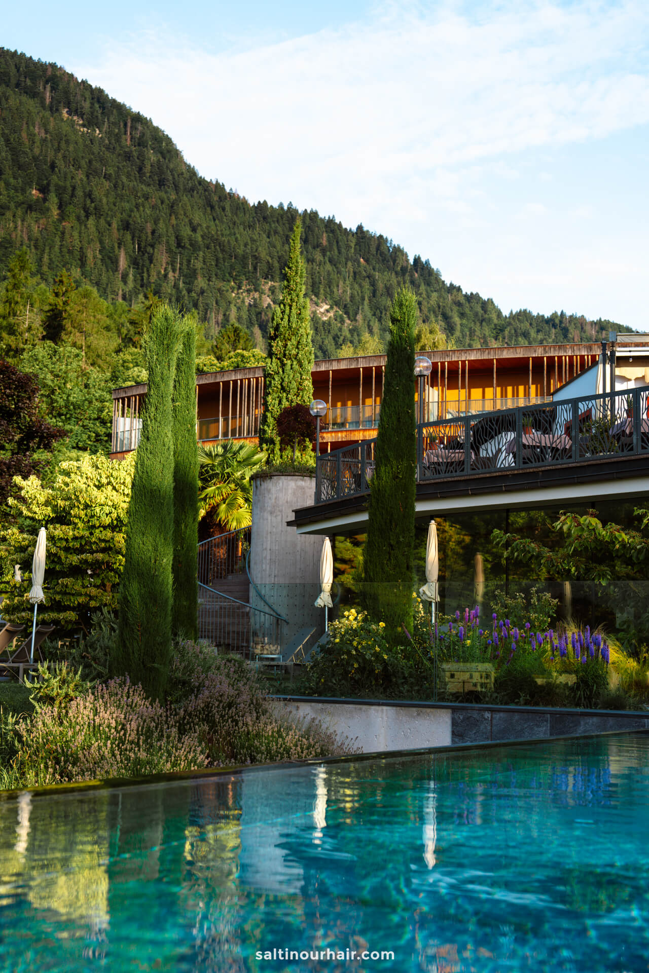 The modern architecture of Hotel Der Waldhof overlooking a landscaped garden with tall trees and a clear swimming pool, set against Merano Dolomites Italy forested mountain backdrop.