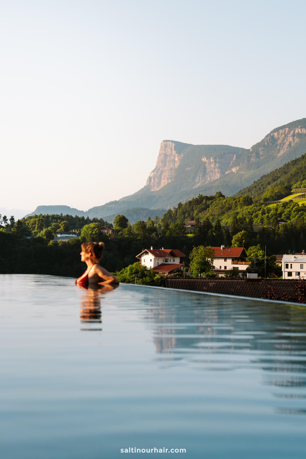 A woman stands in the infinity pool of Hotel Der Waldhof overlooking the village of Merano Italy, with charming houses, lush trees, and mountains in the background.