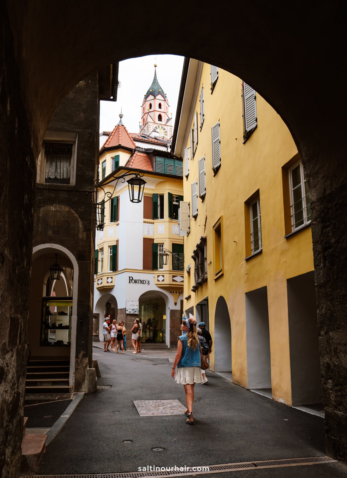 A woman walks through a narrow, arched passageway in Merano Italy with colorful buildings and a clock tower in the background.