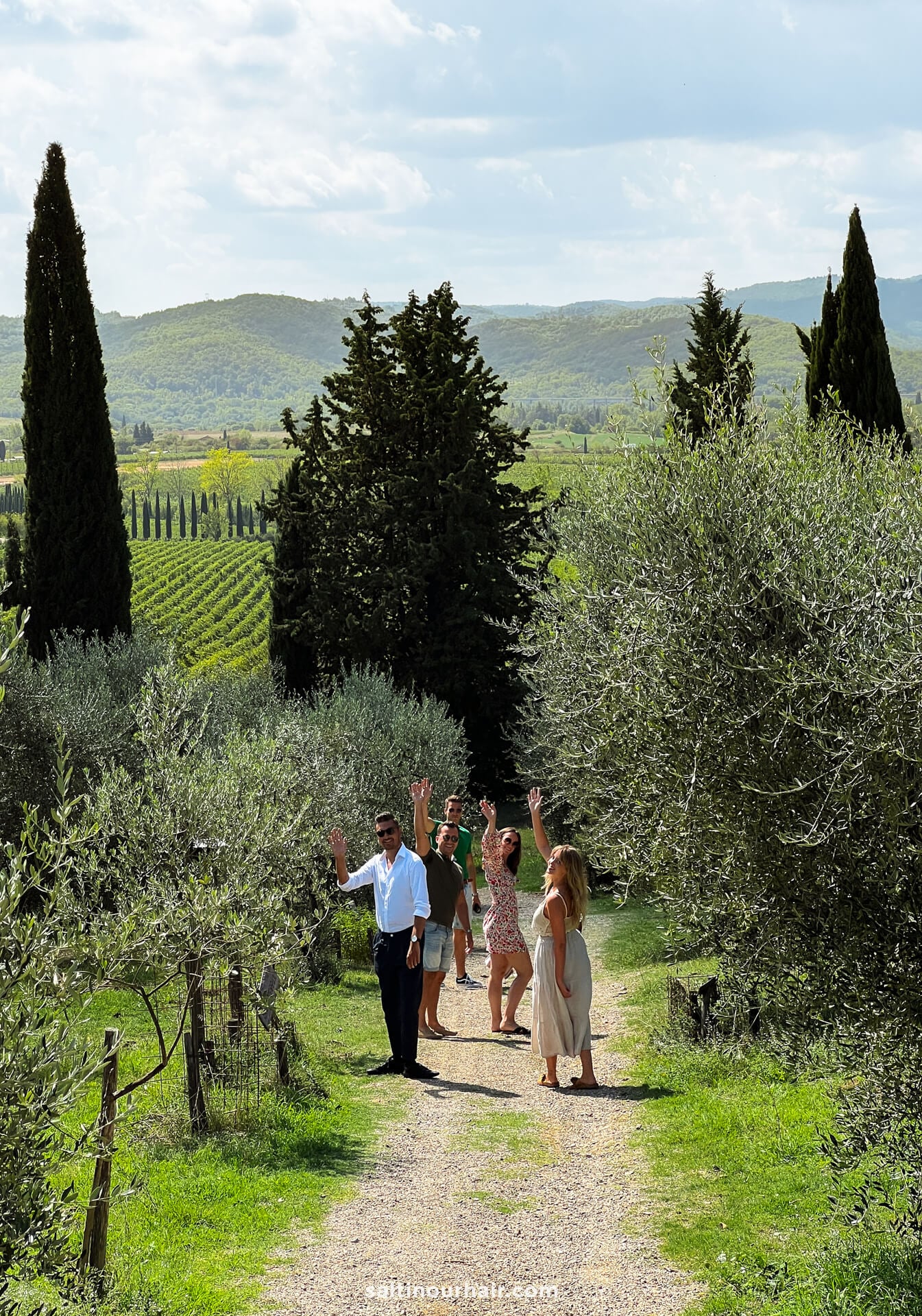 Four people walk along a dirt path between trees on a Tuscany farm in the green countryside, wondering around in the vineyards.