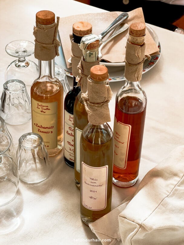 Five glass bottles of oil and vinegar from Fattoria la Vialla in Tuscany Italy, each with cork tops and handwritten labels, are arranged on a set dining table with plates and glasses.