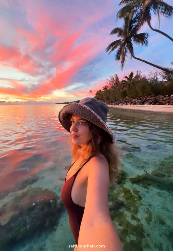 A woman in a hat takes a selfie while standing in clear, shallow water near a tropical beach at sunset with pink clouds in the sky&mdash;one of the unforgettable things to do Rarotonga has to offer.