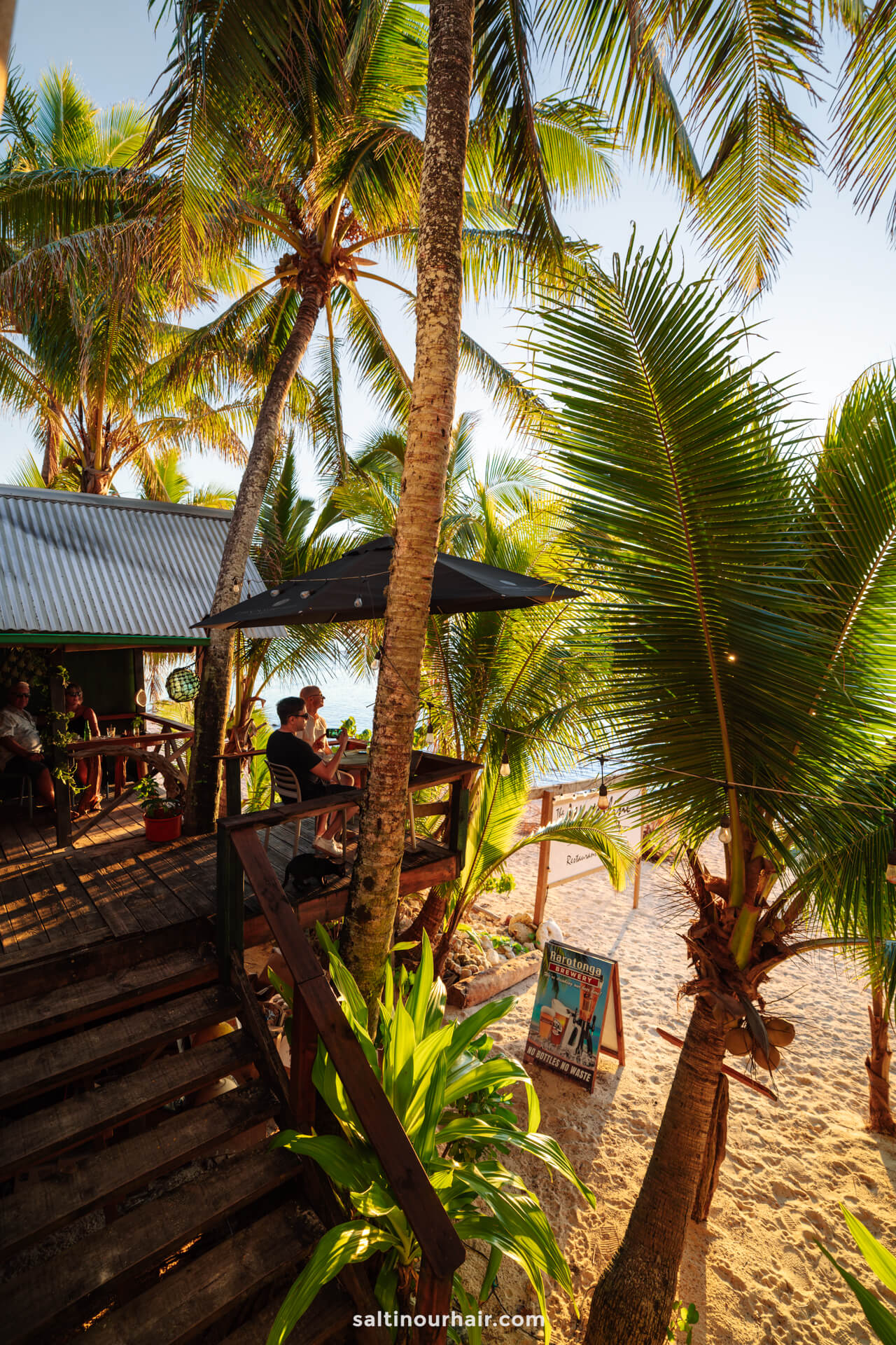 Outdoor beachside caf&eacute; with wooden deck and tables, surrounded by palm trees and plants, overlooking a sunny sandy beach. A man sits at a table near a signboard&mdash;a relaxing spot after exploring things to do in Rarotonga, Cook Islands