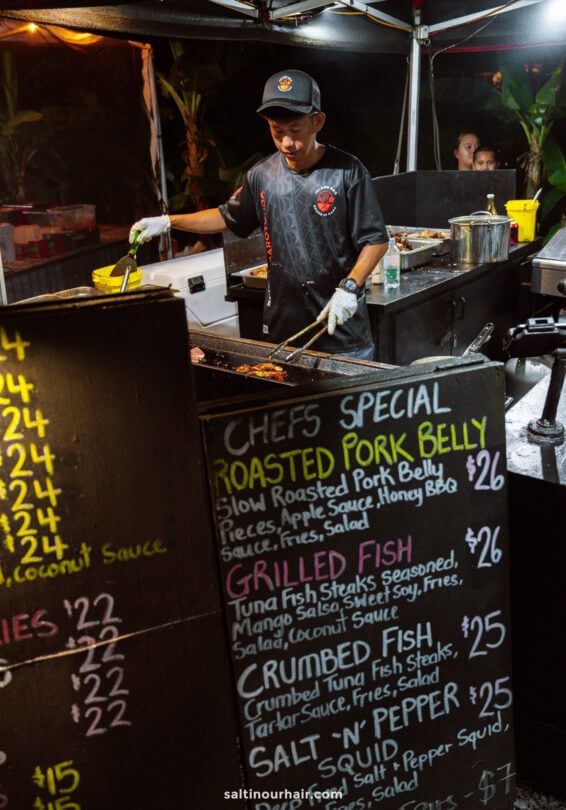 A chef grills meat at a night market food stall, with a chalkboard menu listing specials like roasted pork belly and grilled fish&mdash;one of the top things to do in Rarotonga for food lovers.