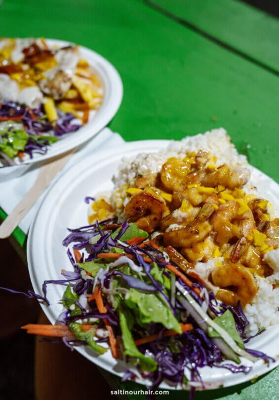 Two plates of food featuring grilled shrimp with sauce over rice, served with mixed salad greens and shredded vegetables, on a green table&mdash;a delicious highlight among the things to do in Rarotonga.