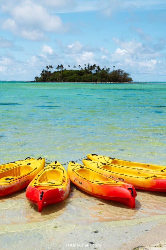 Five yellow and red kayaks rest on a sandy shore with clear blue-green water and a tree-covered island in the distance&mdash;a perfect glimpse of the many things to do in Rarotonga, Cook Islands