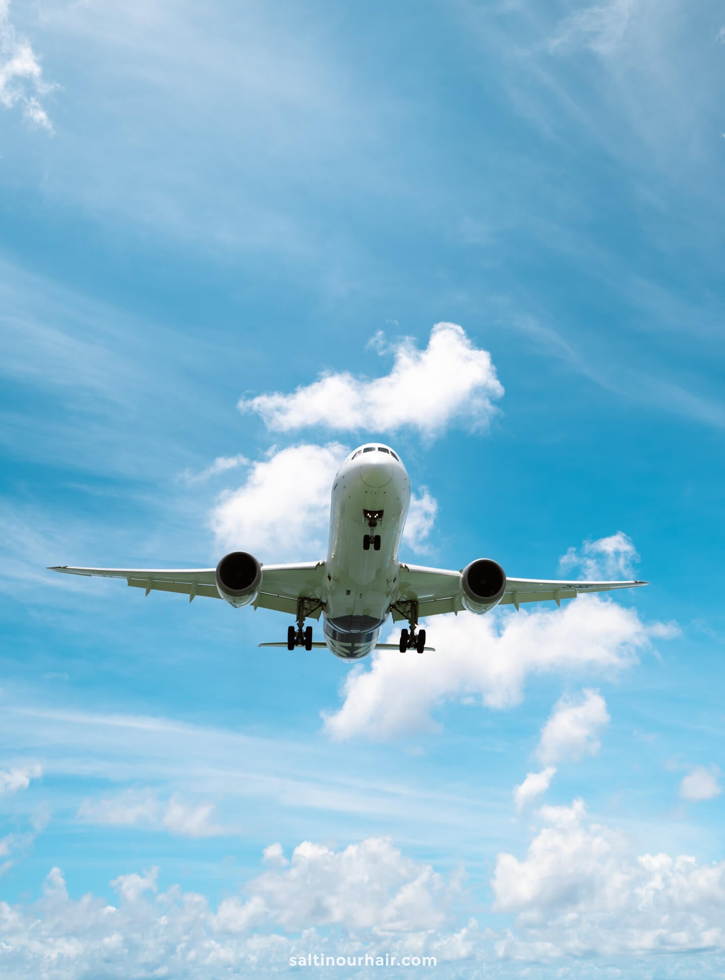 A commercial airplane flying against a bright blue sky with scattered white clouds, viewed from below&mdash;reminding you of the incredible things to do in Rarotonga, Cook Islands
