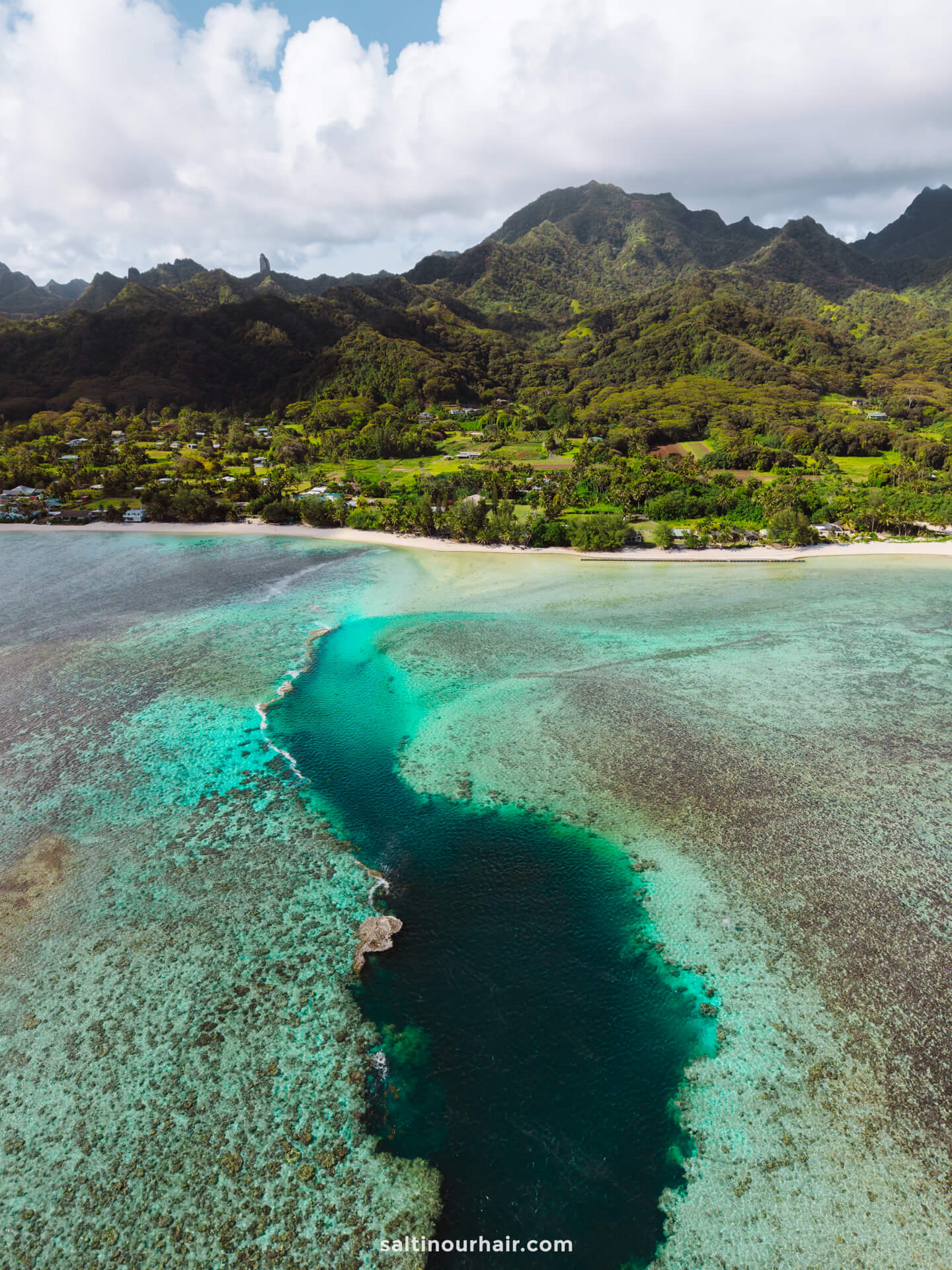 A coastal landscape with a turquoise lagoon and coral reef, bordered by green mountains and a cloudy sky in the background&mdash;perfect for discovering the best things to do Rarotonga has to offer.