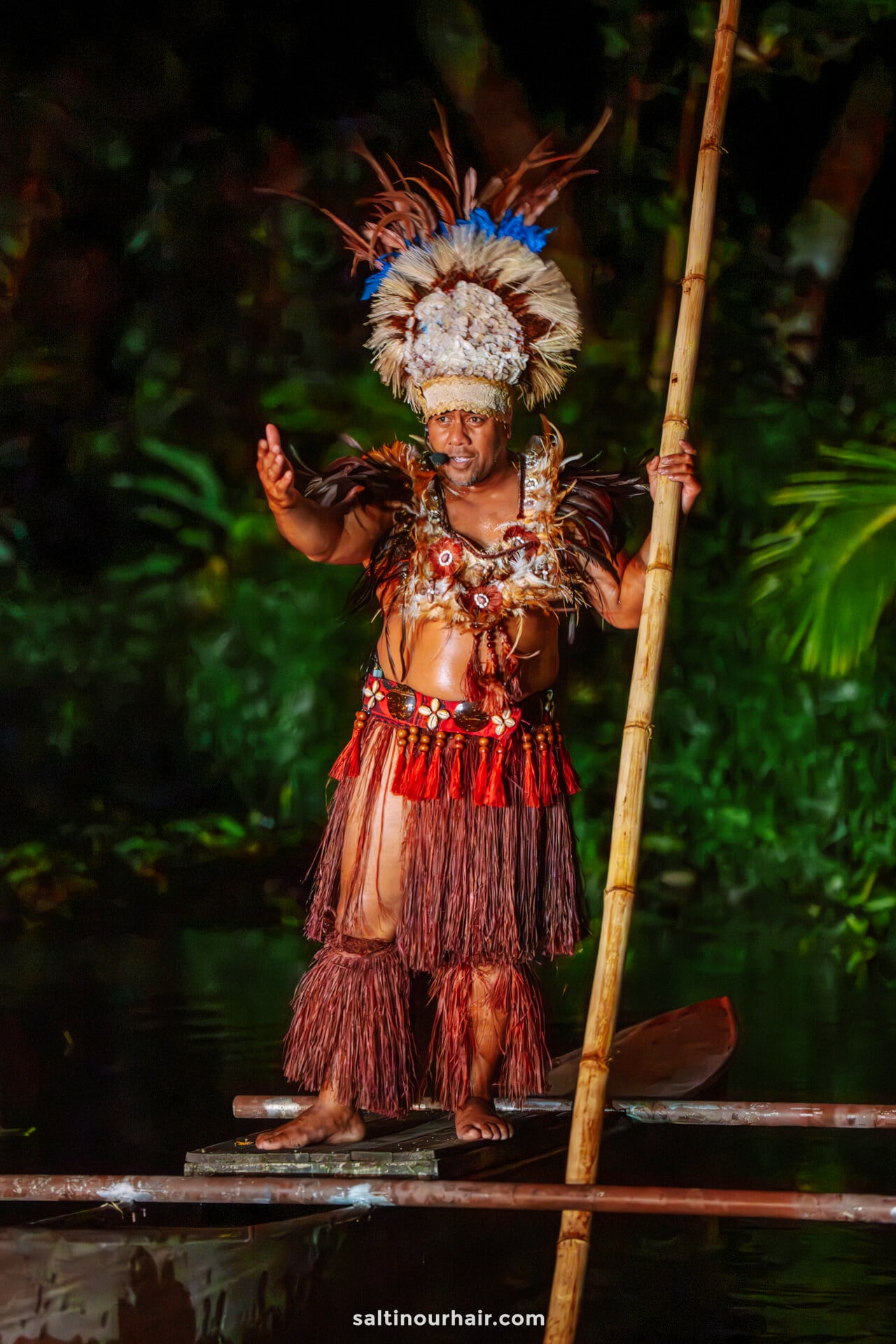 A person wearing traditional Polynesian attire and headdress stands on a wooden raft, holding a long pole, with lush greenery in the background&mdash;capturing the authentic spirit of things to do Rarotonga offers visitors.