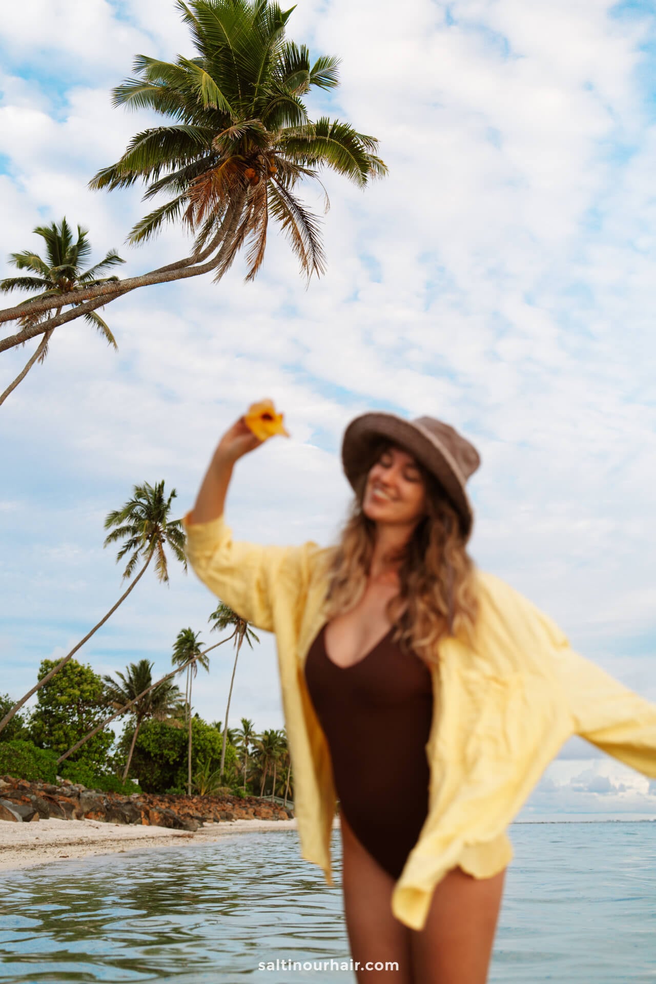 A woman in a brown swimsuit and yellow shirt stands in shallow water on a tropical beach, holding a yellow flower&mdash;one of the idyllic things to do Rarotonga offers, with palm trees and a partly cloudy sky in the background.