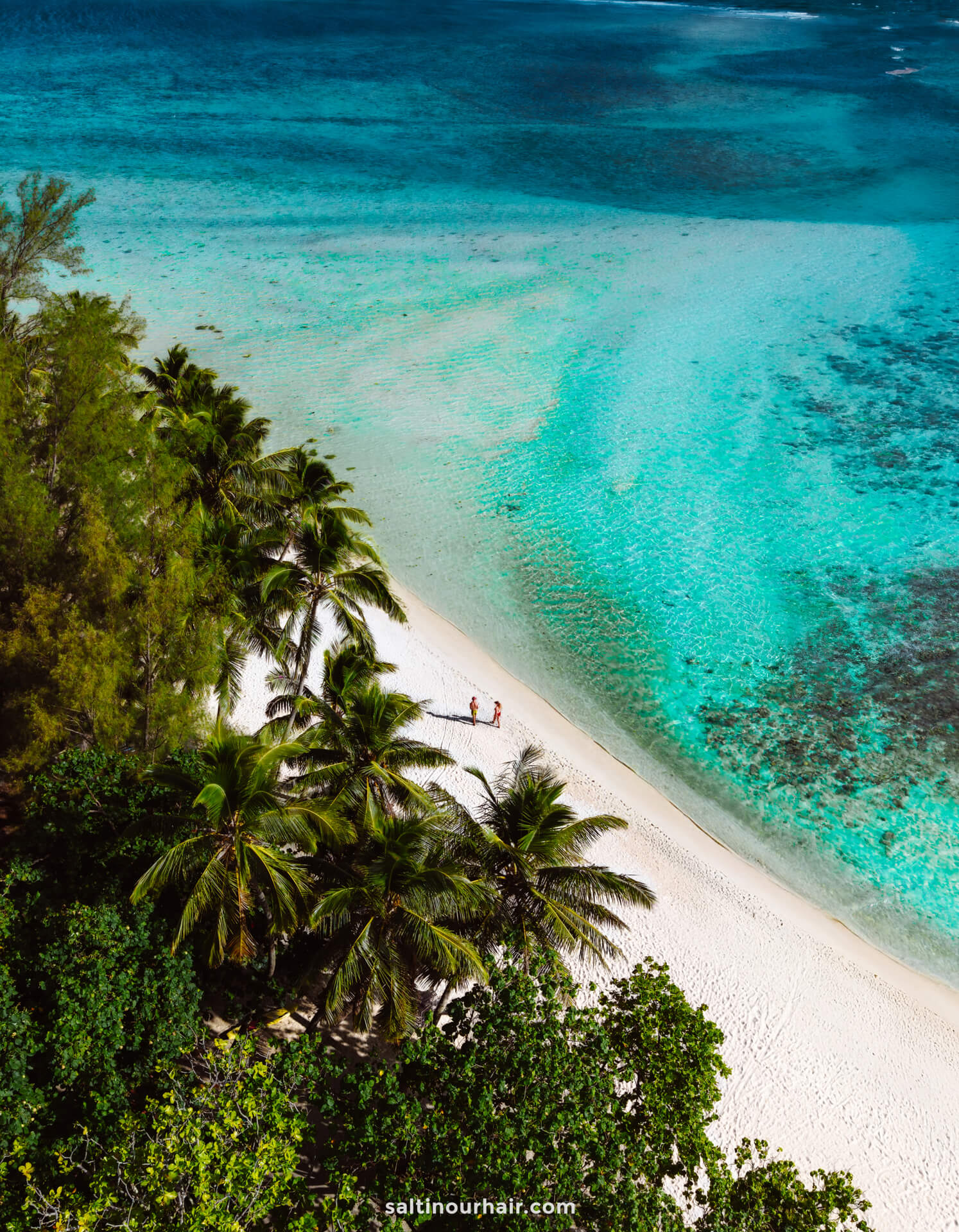 Aerial view of a tropical beach with white sand, turquoise water, palm trees, and two people walking along the shoreline&mdash;a perfect spot for your list of things to do Rarotonga, Cook Islands