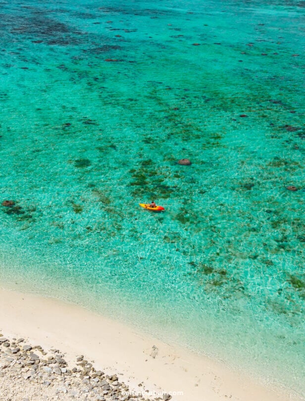 A person in an orange kayak paddles over clear turquoise water near a sandy beach with visible rocks and shallow areas&mdash;one of the top things to do in Rarotonga, Cook Islands