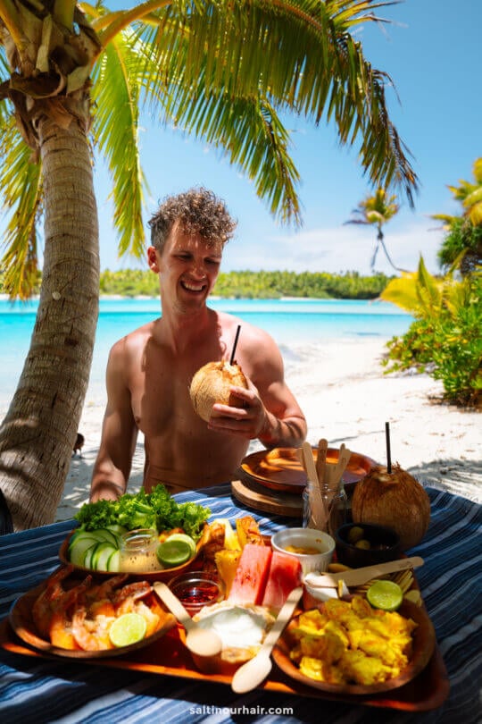 A shirtless person sits at a tropical beachside table in Rarotonga, holding a coconut drink, with assorted fruit and food dishes in front of them under palm trees&mdash;one of the top things to do in Rarotonga, Cook Islands