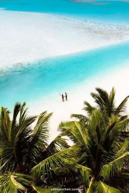 Two people stand on a white sandy beach near turquoise water, surrounded by tropical palm trees under a clear sky&mdash;one of the unforgettable things to do in Rarotonga, Cook Islands