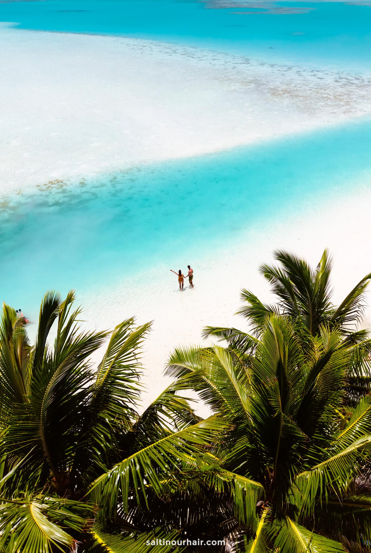 Two people stand on a white sandy beach near turquoise water, surrounded by tropical palm trees under a clear sky&mdash;one of the unforgettable things to do in aitutaki, cook islands.
