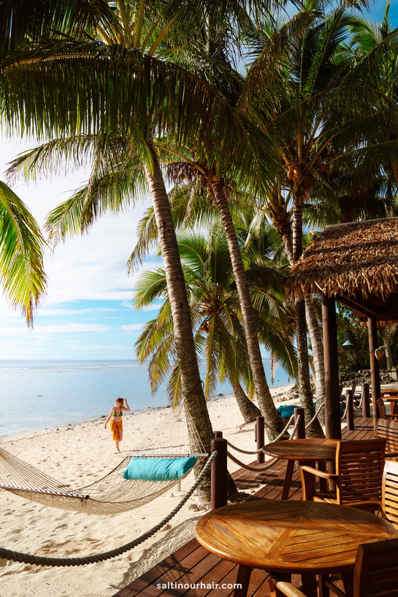 A tropical beach scene with palm trees, a hammock, wooden chairs and tables on a deck, and a person walking on the sand near the shoreline&mdash;perfect for relaxing or discovering things to do in Rarotonga, Cook Islands