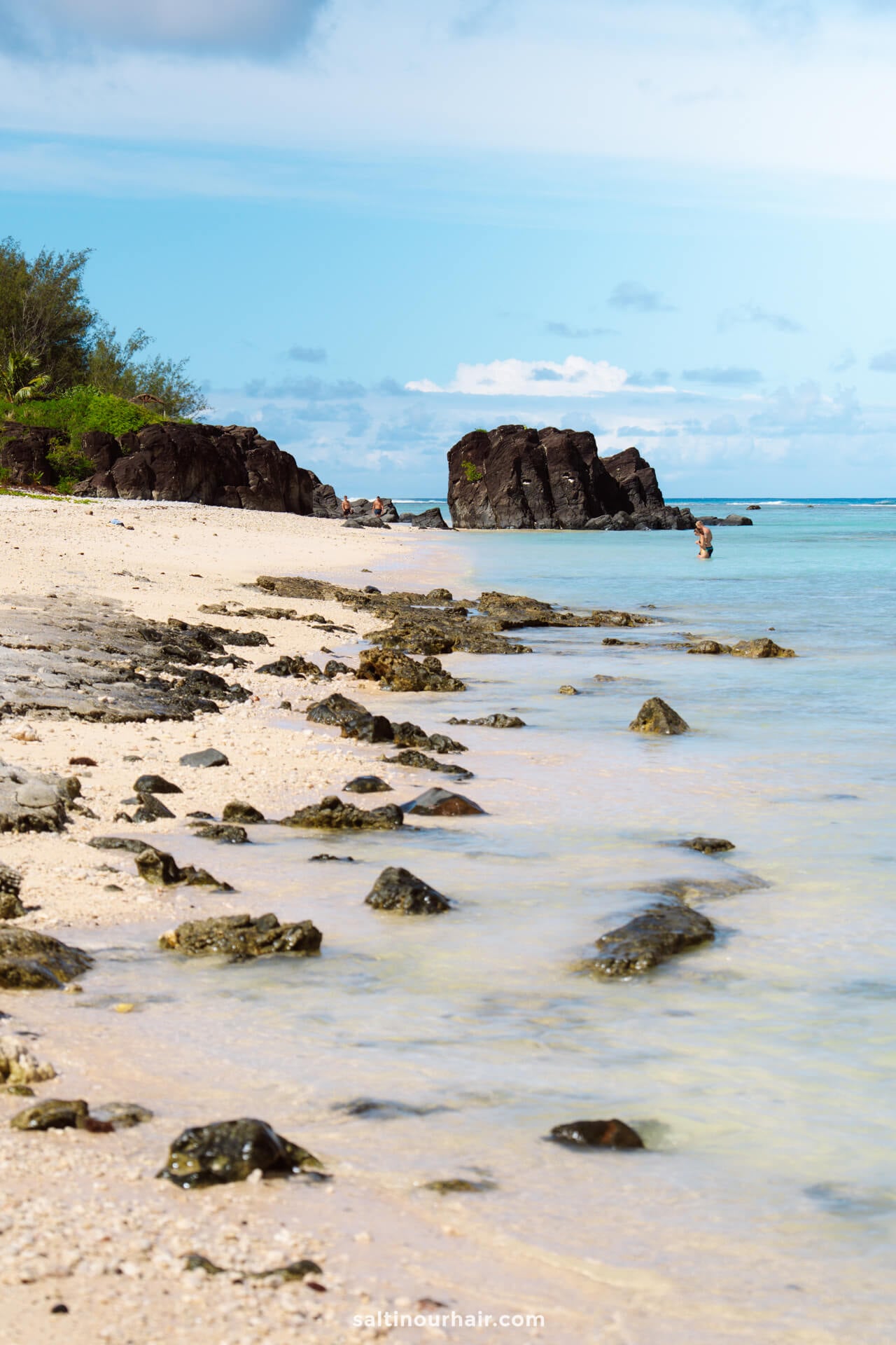 A sandy beach with scattered rocks leads to turquoise water; dark rock formations and sparse greenery are visible in the background under a partly cloudy sky, offering a peaceful spot among the many things to do in Rarotonga, Cook Islands