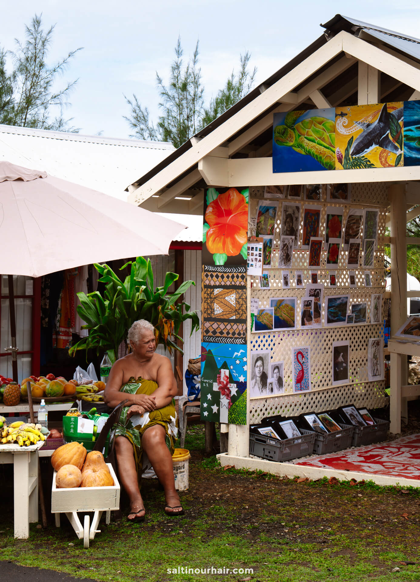 An elderly woman sits beside a table of fruits outside a small art shop displaying colorful paintings and prints&mdash;a charming glimpse into local life among the many things to do in Rarotonga, Cook Islands