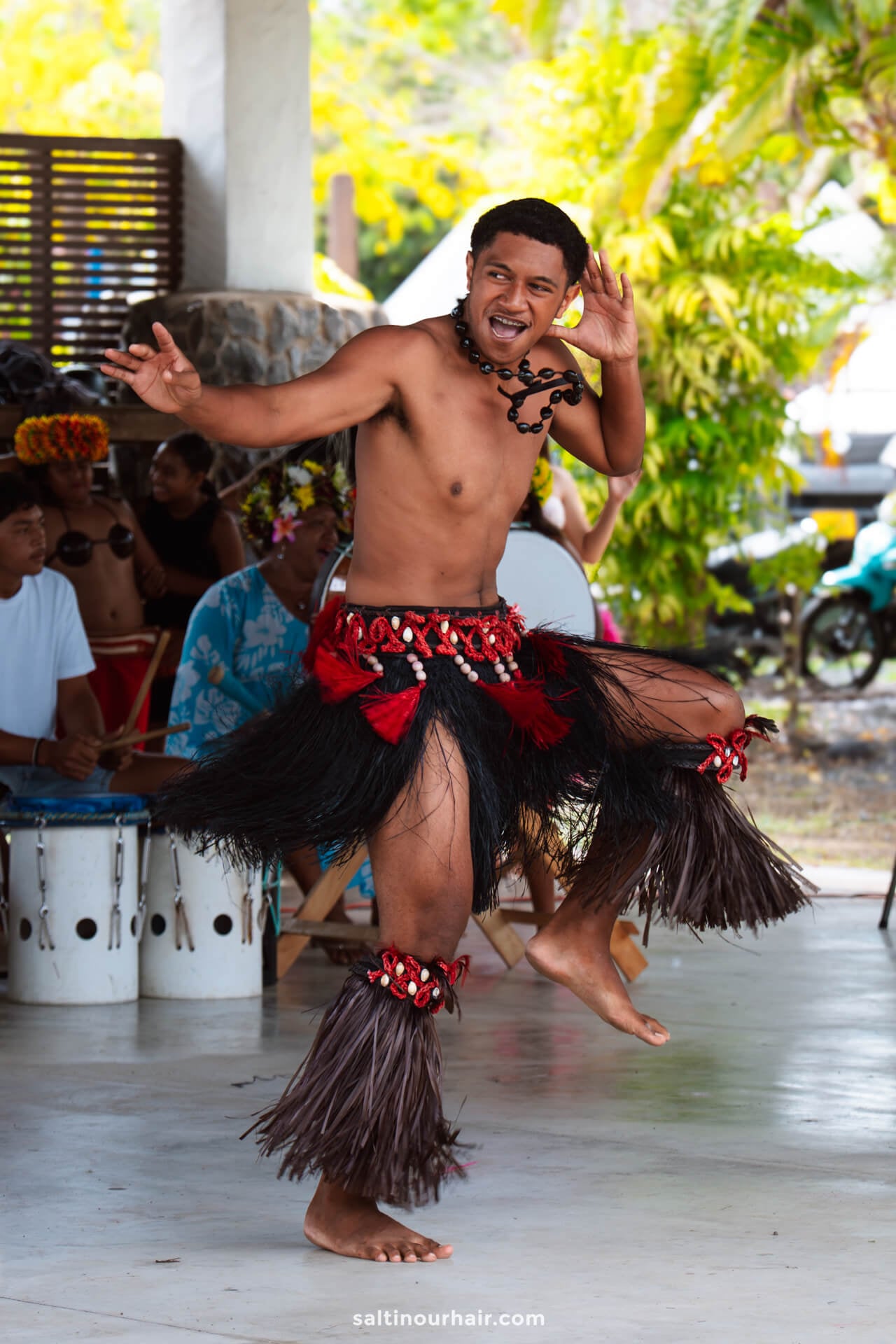 A man in traditional Polynesian attire performs a dance on a covered outdoor stage, with musicians playing drums in the background&mdash;one of the vibrant things to do Rarotonga offers visitors.