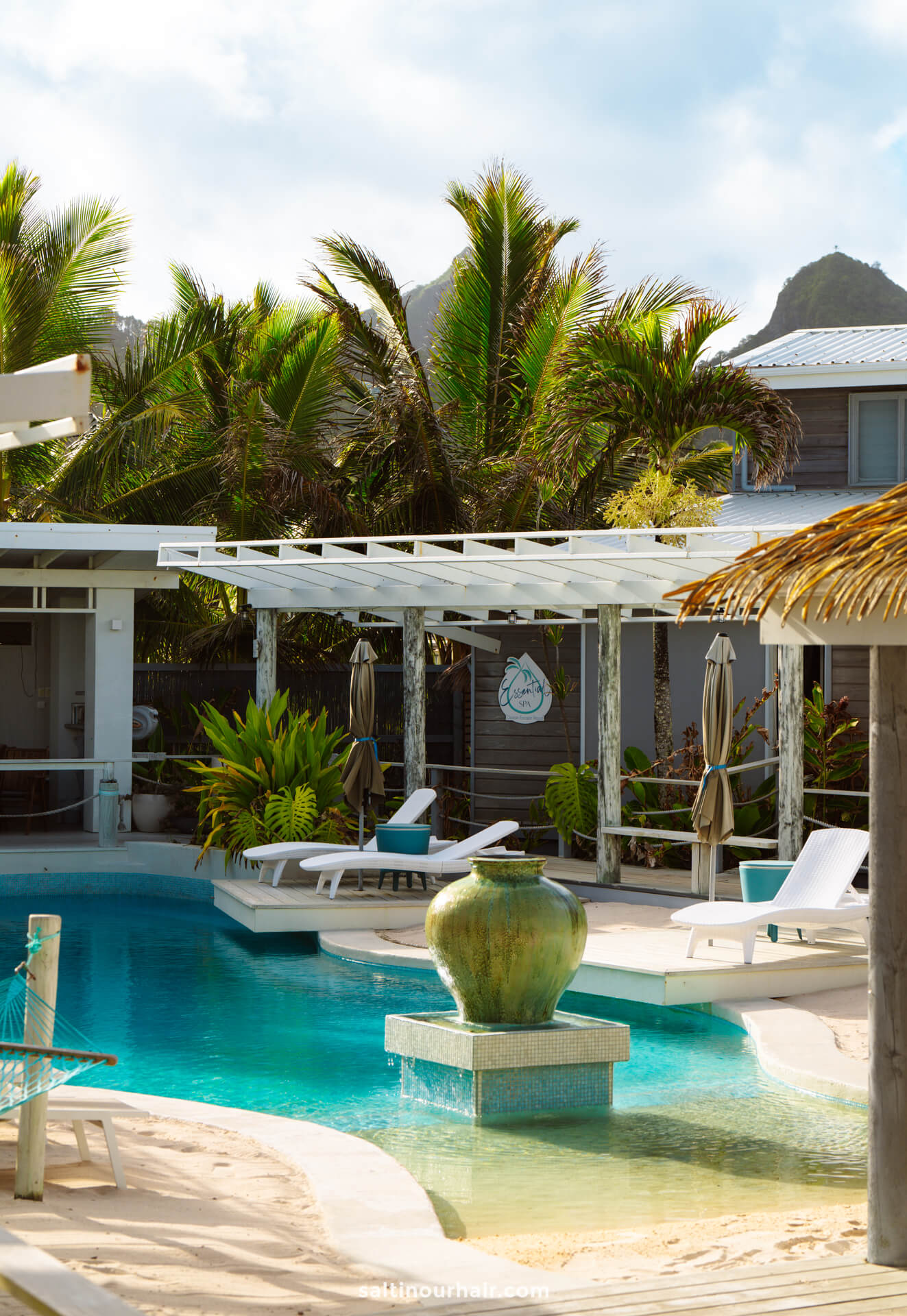 A pool area with a large ceramic fountain in the center, surrounded by lounge chairs, palm trees, and a pergola&mdash;perfect for relaxing after exploring the many things to do in Rarotonga at this tropical resort setting.