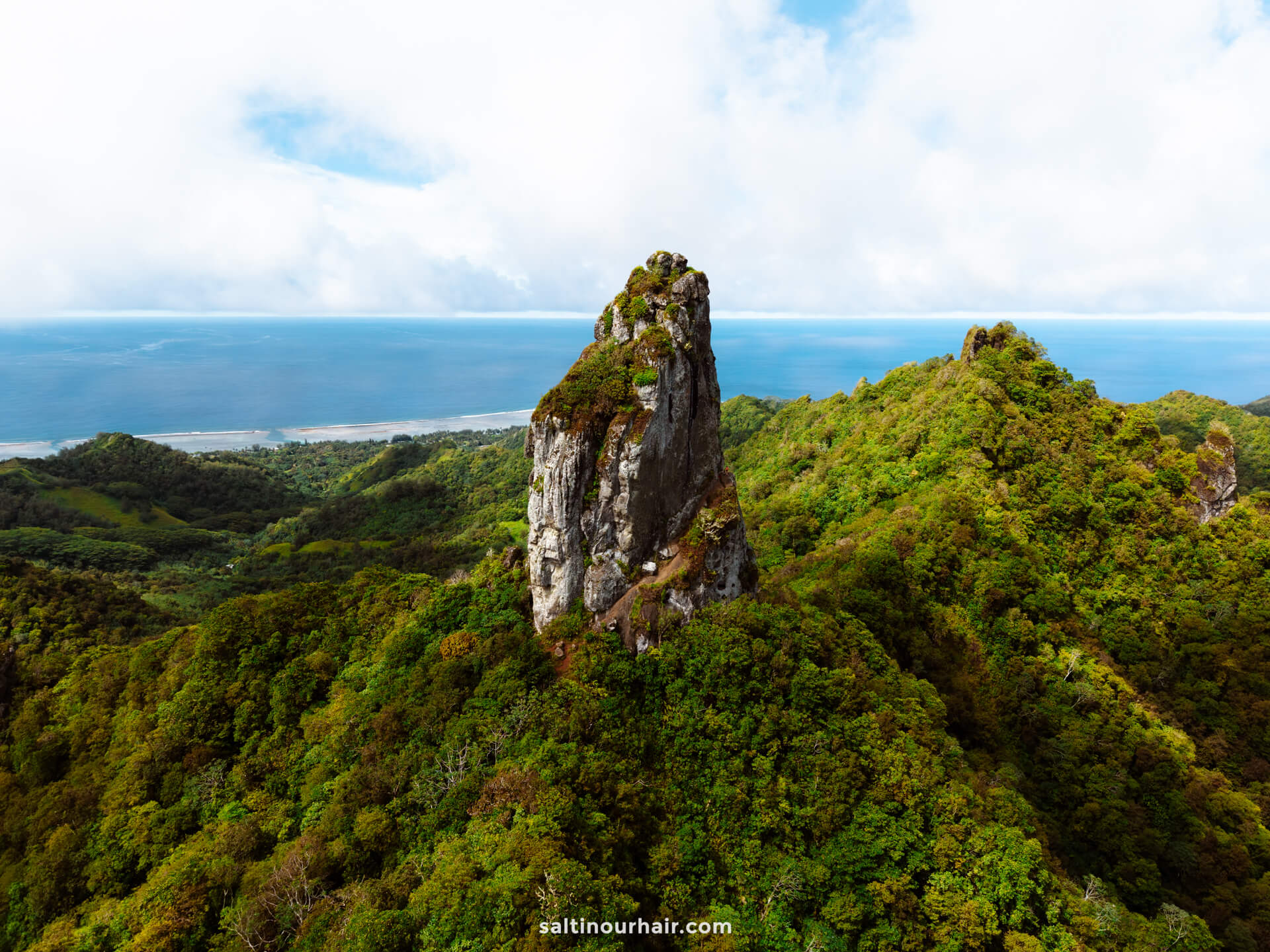 A tall, rocky peak rises above dense green forest with ocean visible in the distance under a partly cloudy sky&mdash;one of the breathtaking sights among the best things to do in Rarotonga, Cook Islands