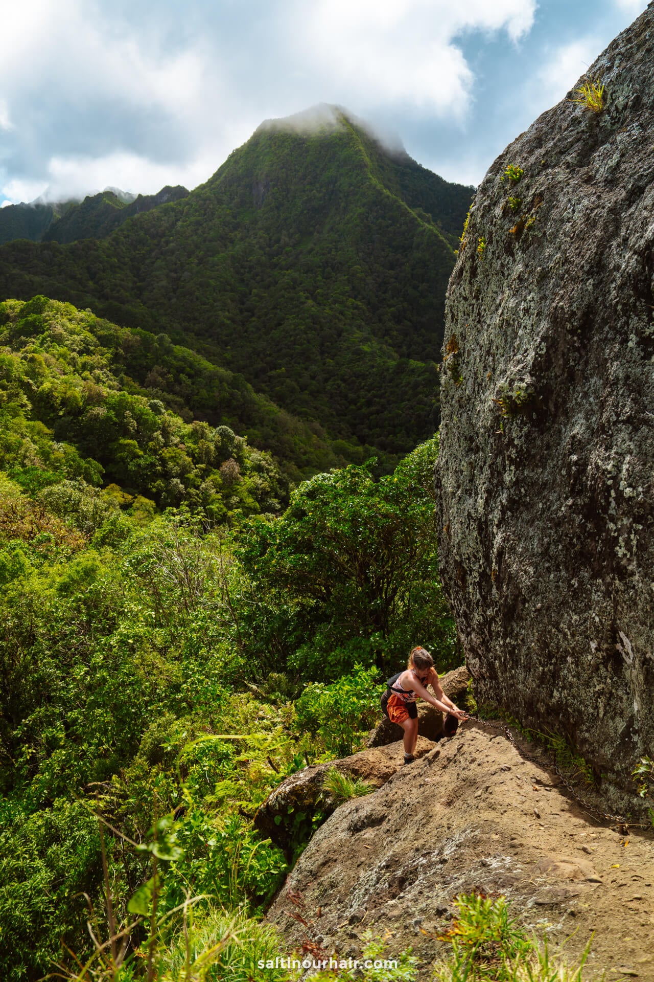 A person climbs a rocky trail beside a large boulder, with lush green mountains and a cloudy sky in the background&mdash;one of the adventurous things to do Rarotonga offers.