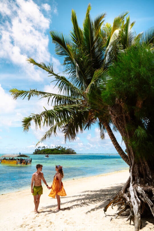 A man and woman holding hands stand on a sandy beach under palm trees in Rarotonga, with clear blue water and a small boat in the background&mdash;one of the idyllic things to do in Rarotonga, Cook Islands